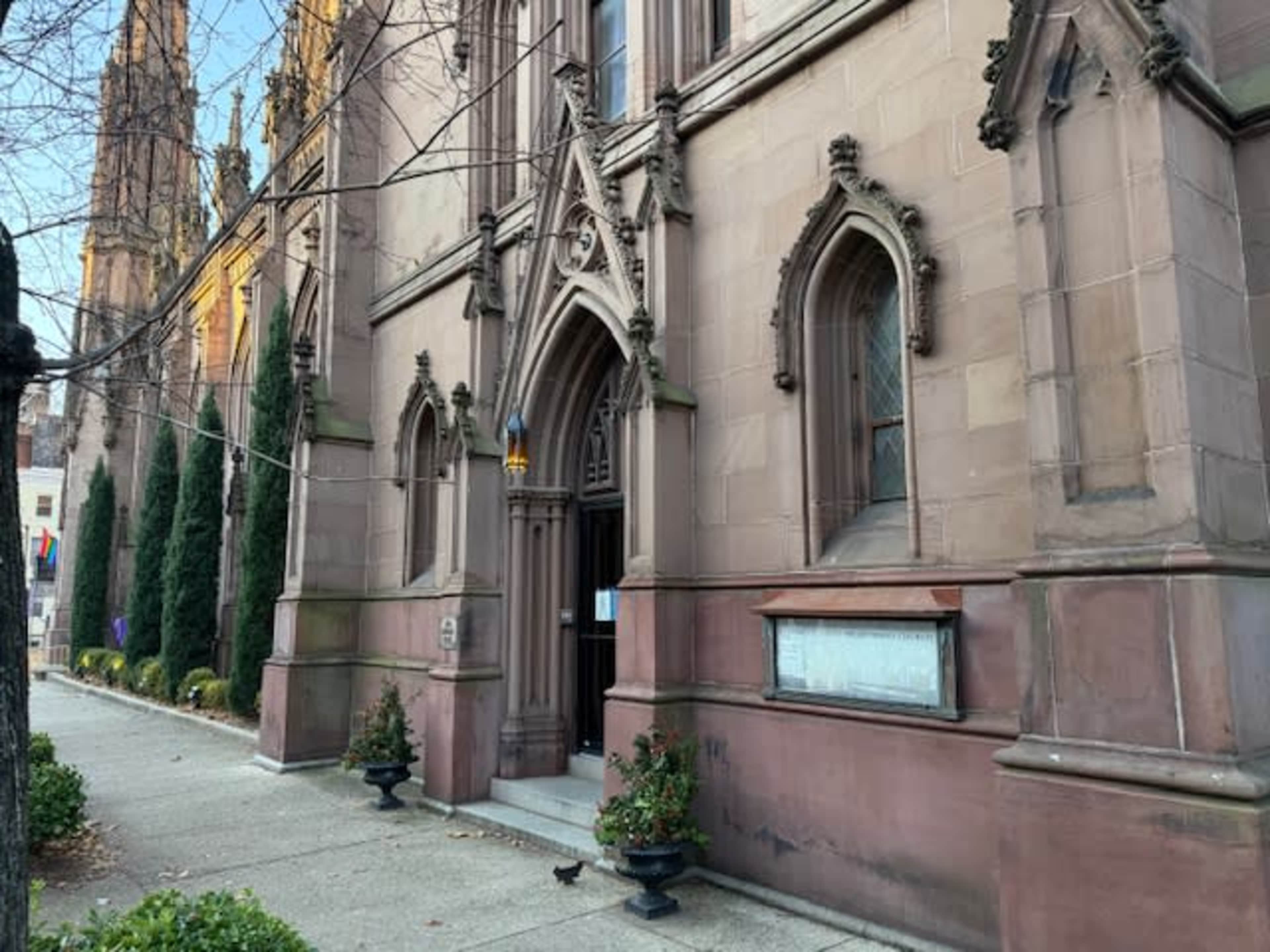 The image shows the entrance of a historic stone building with gothic architectural features, framed by neatly trimmed shrubs and small planters.