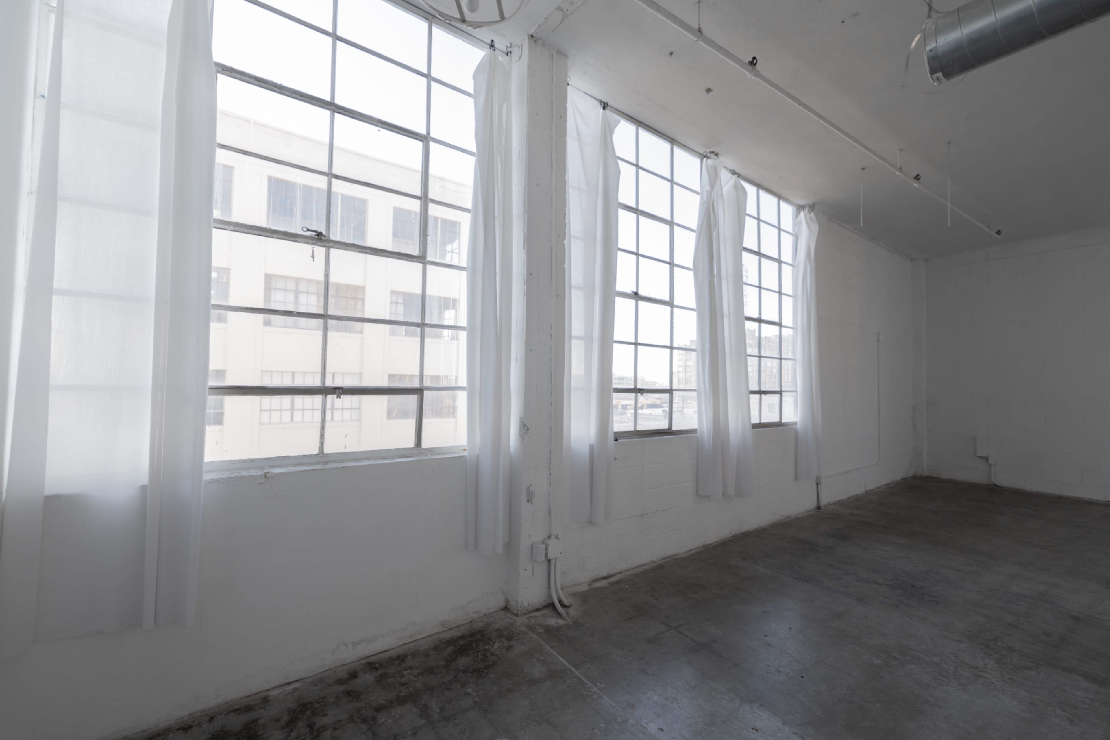 The image shows a minimalist, empty room with large windows dressed in sheer white curtains, allowing natural light to fill the space.