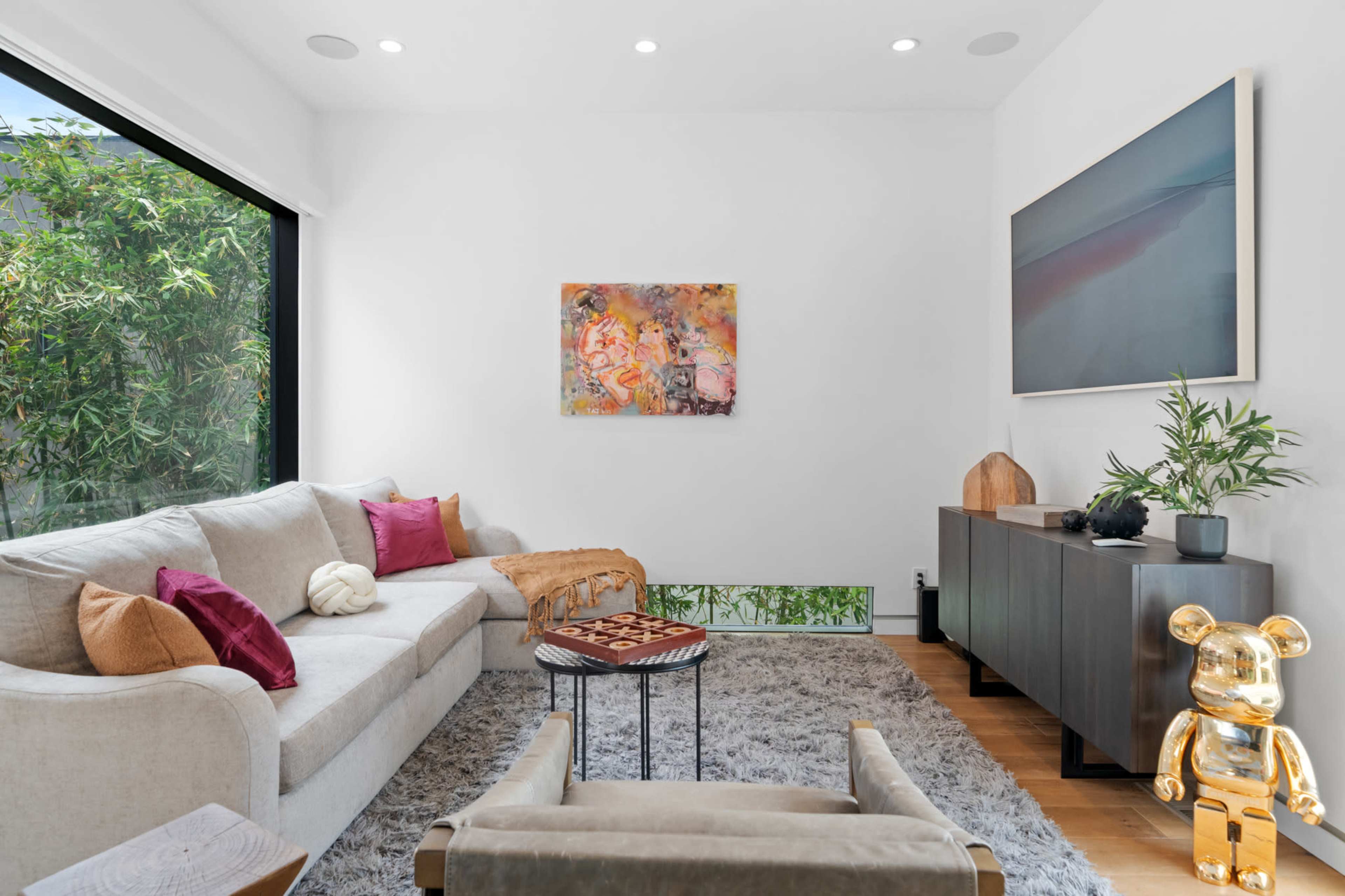 A modern living room features a light-colored sectional sofa, a textured rug, a wooden sideboard, and an art piece hanging on the wall beside a large window.