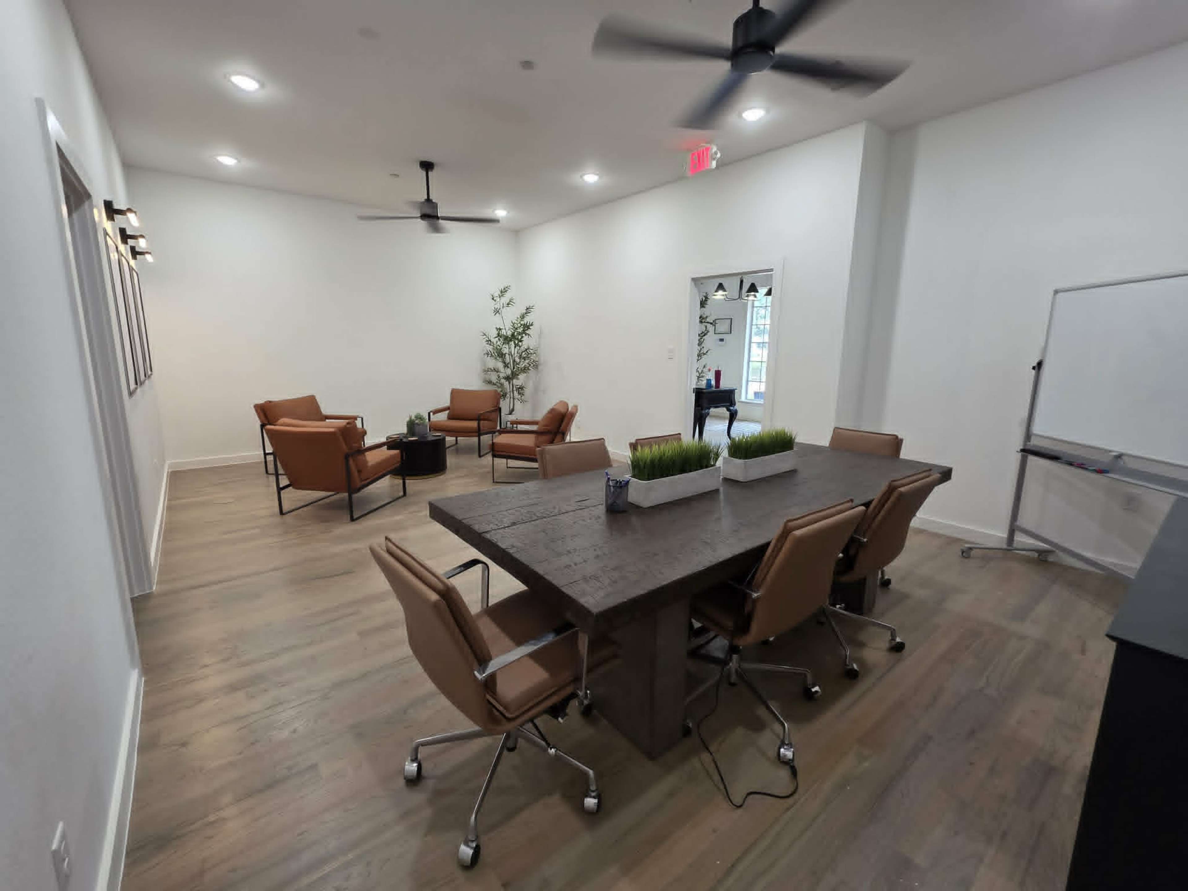 The image shows a modern conference room featuring a large wooden table surrounded by ergonomic chairs, along with several brown leather lounge chairs and a whiteboard in the corner.