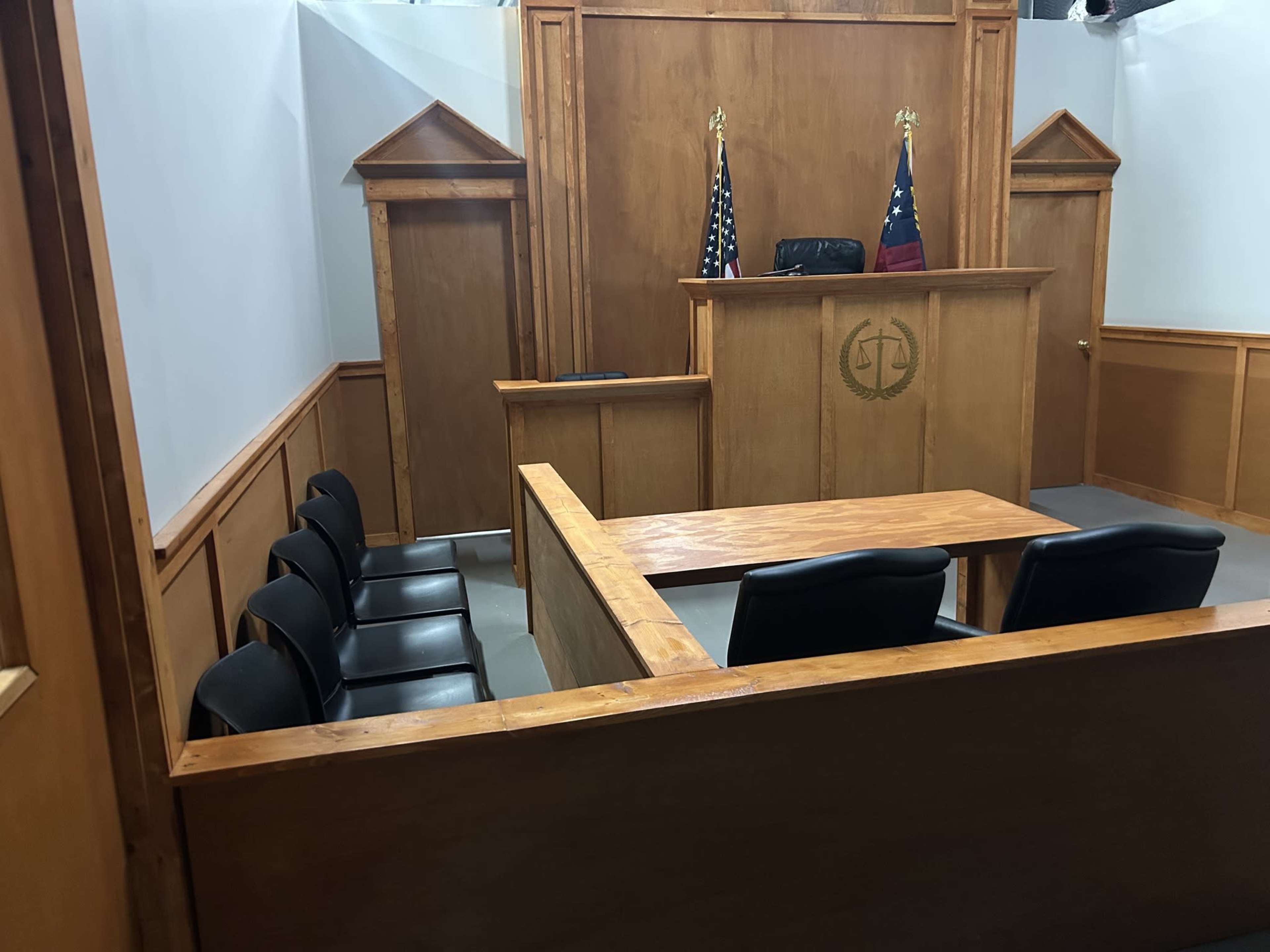 The image depicts a courtroom interior featuring a wooden judge's bench, a jury box with black chairs, and American and state flags.