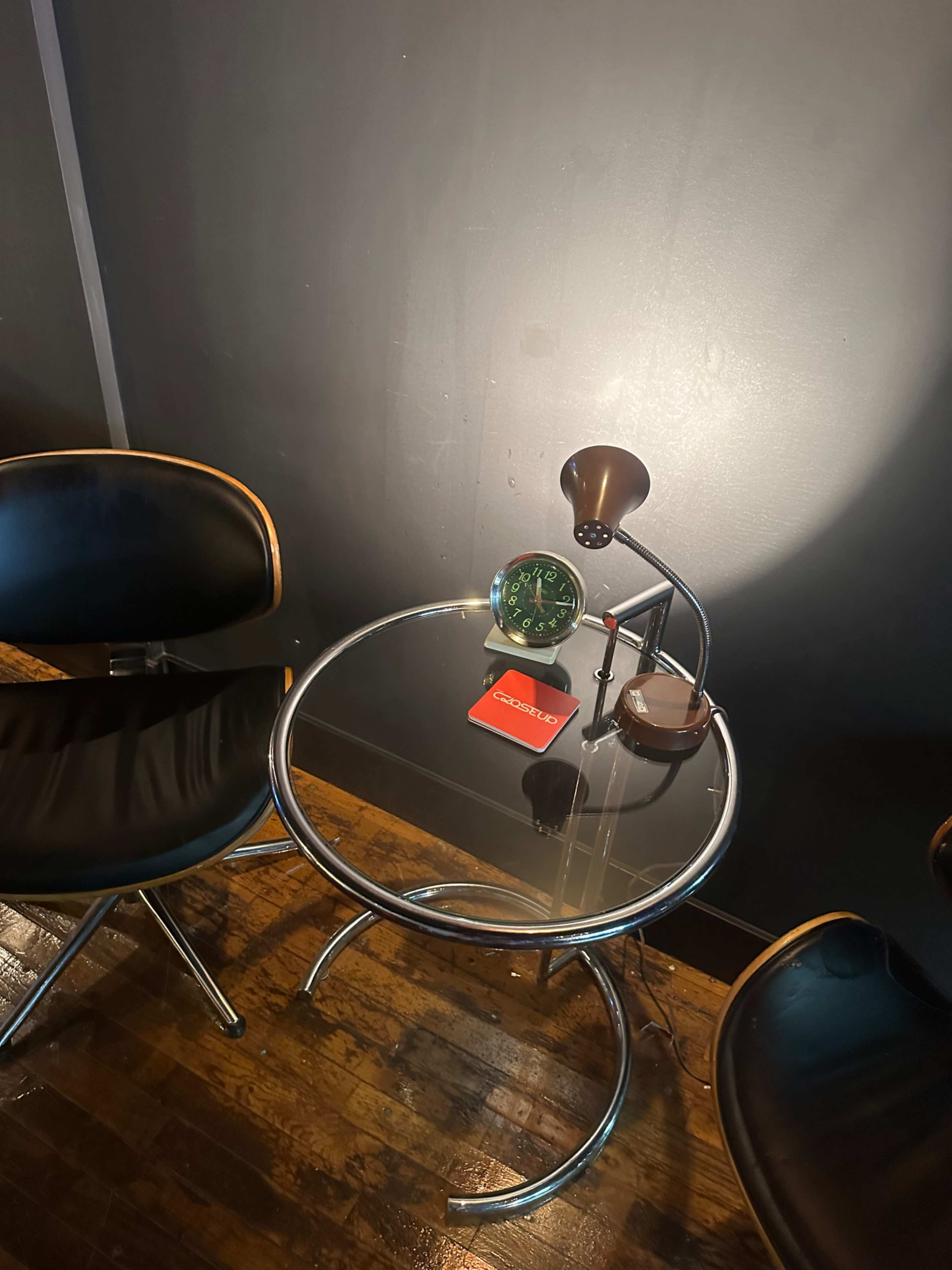 A small glass table with a black lamp and a green decorative item sits between two black chairs against a dark wall.