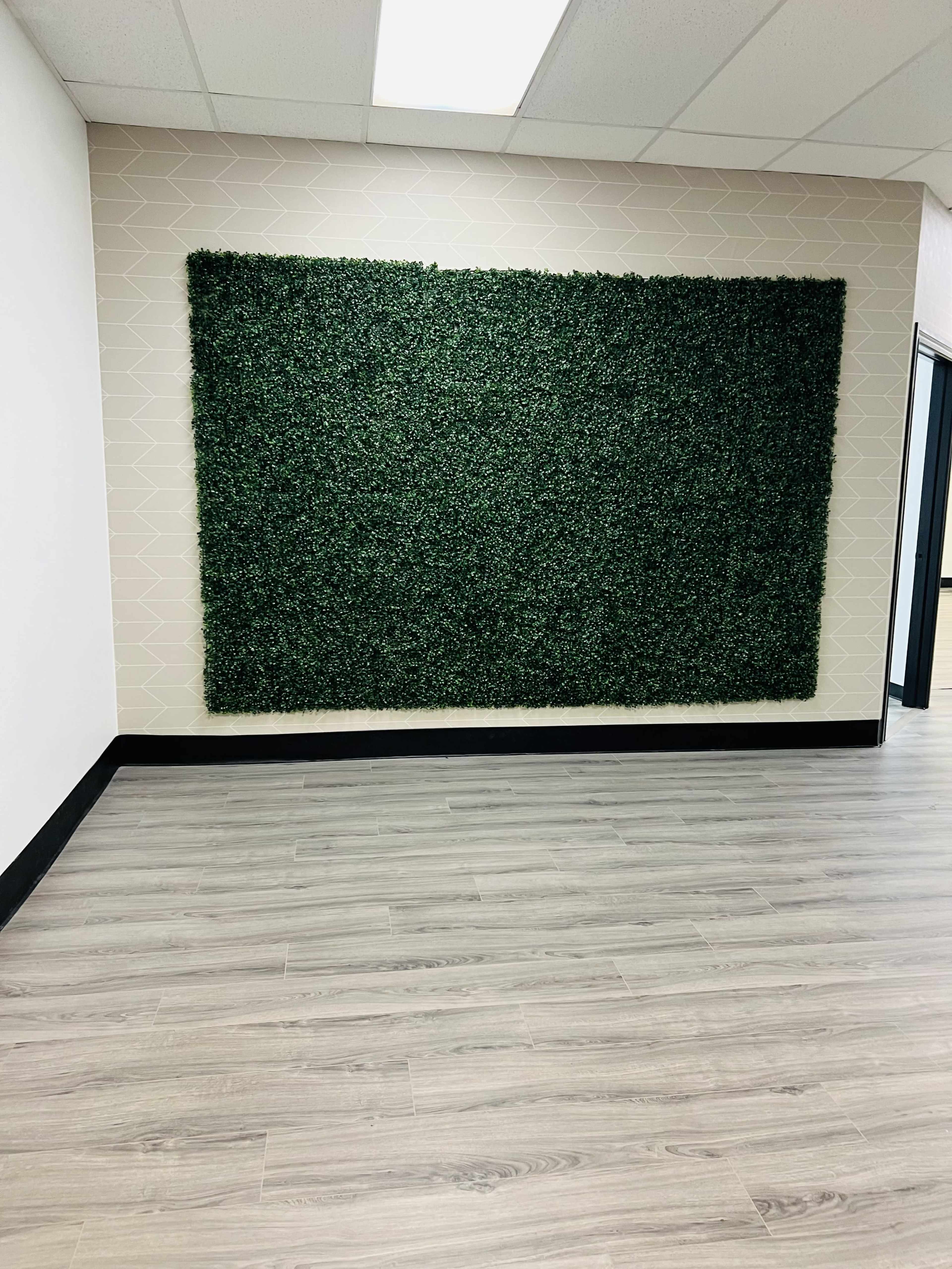 A large green artificial hedge panel mounted on a light-colored wall in a room with wooden flooring.