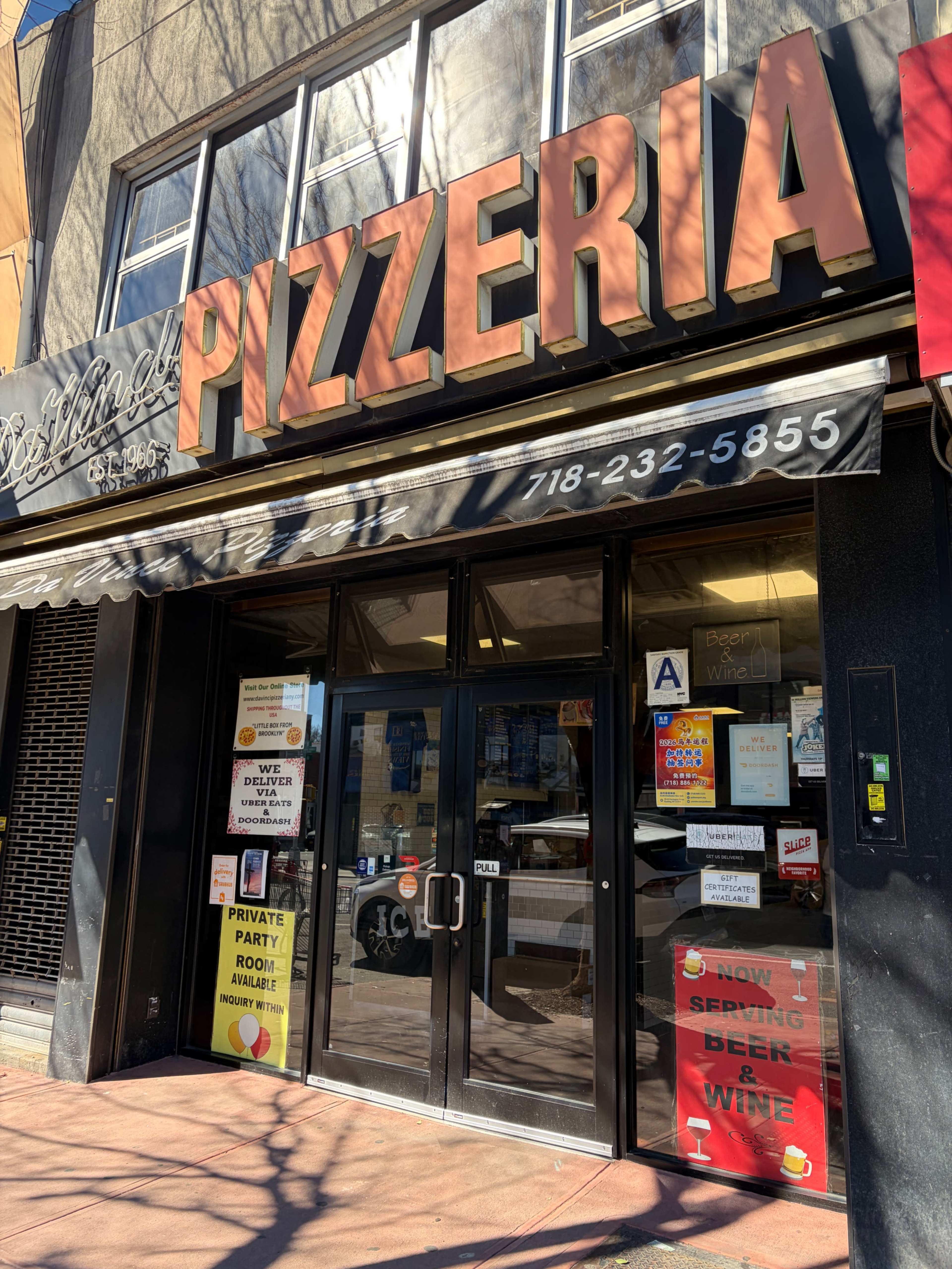 The image shows the storefront of a pizzeria with a large sign above the entrance and various advertisements in the windows, indicating private party options and beer and wine availability.