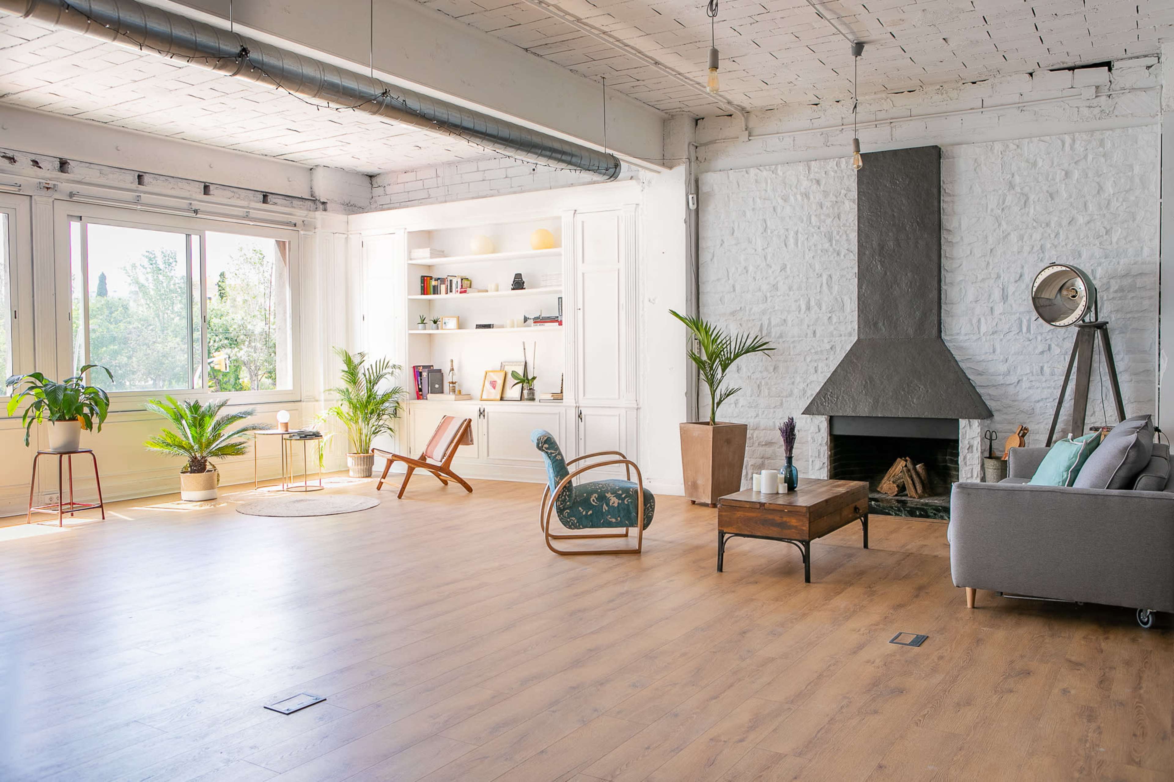 A spacious living area with a large window, a stone fireplace, a wooden floor, and various plants alongside minimalist furniture.
