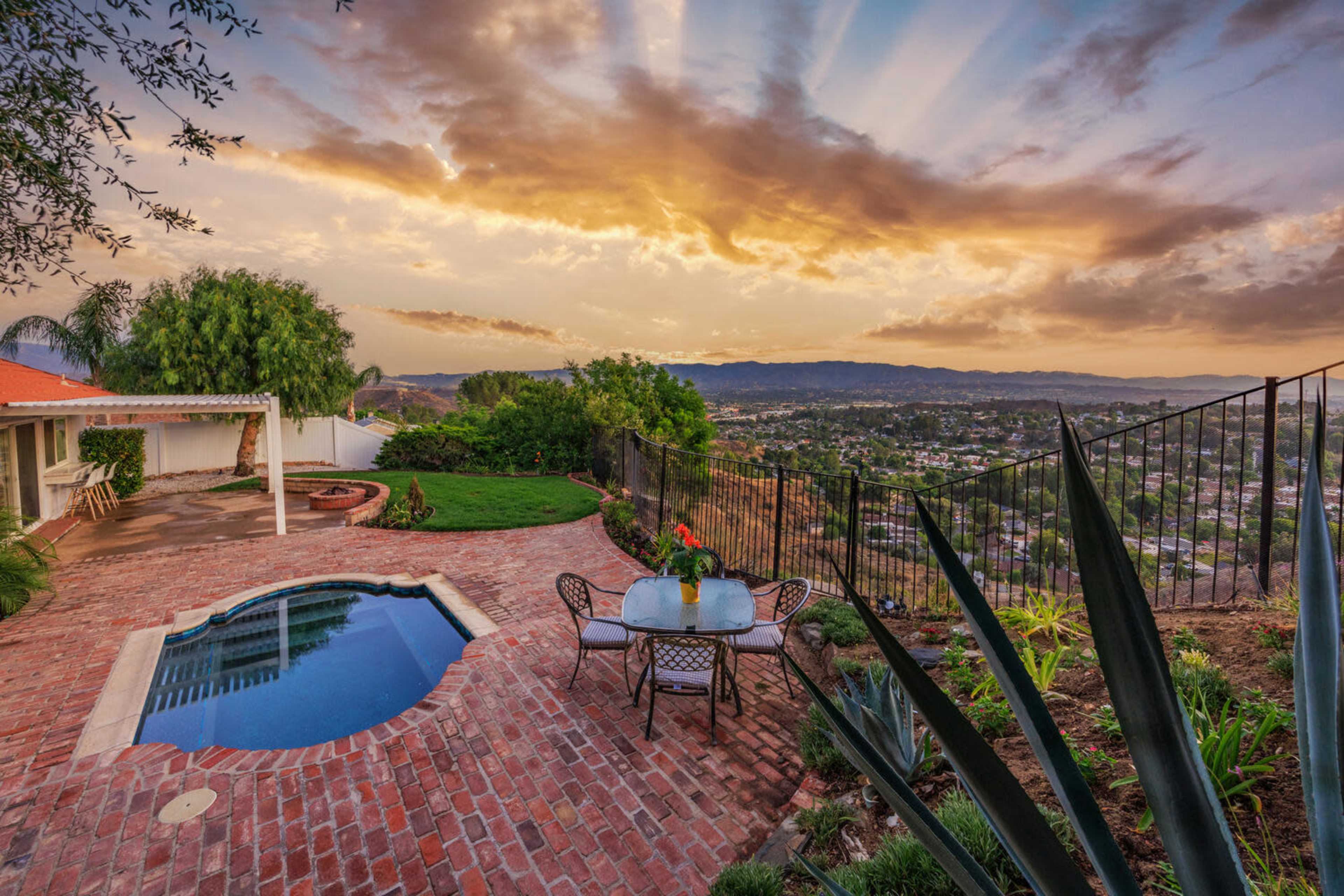 The image shows a backyard with a swimming pool and a small table with chairs, overlooking a valley at sunset.