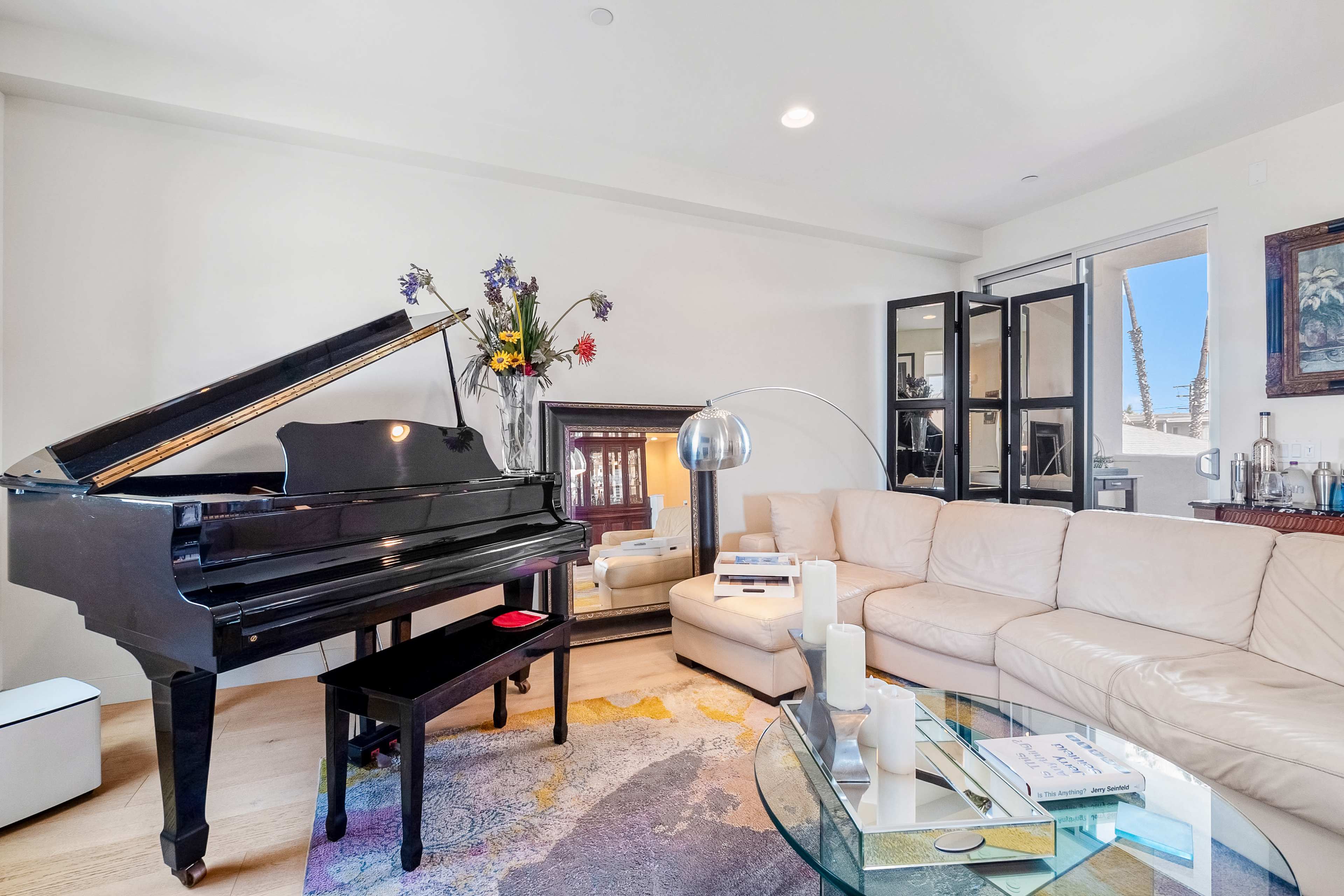 The living room features a grand piano beside a white sectional sofa, a glass coffee table, and large windows with black-frame doors leading outside.