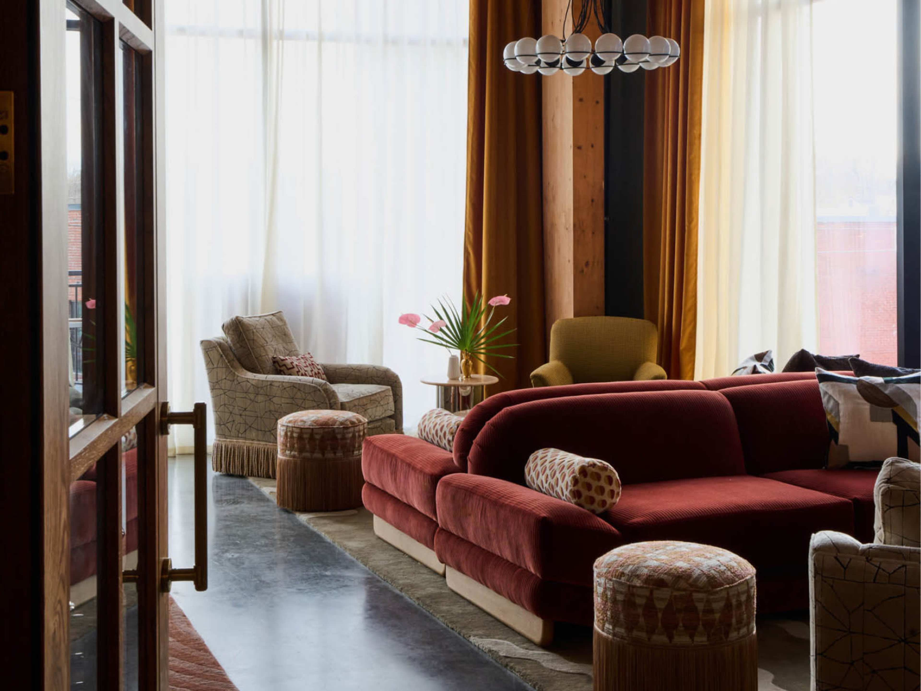 The image depicts a modern living room with a deep red sectional sofa, two patterned armchairs, and decorative stools, surrounded by large windows with sheer curtains letting in natural light.