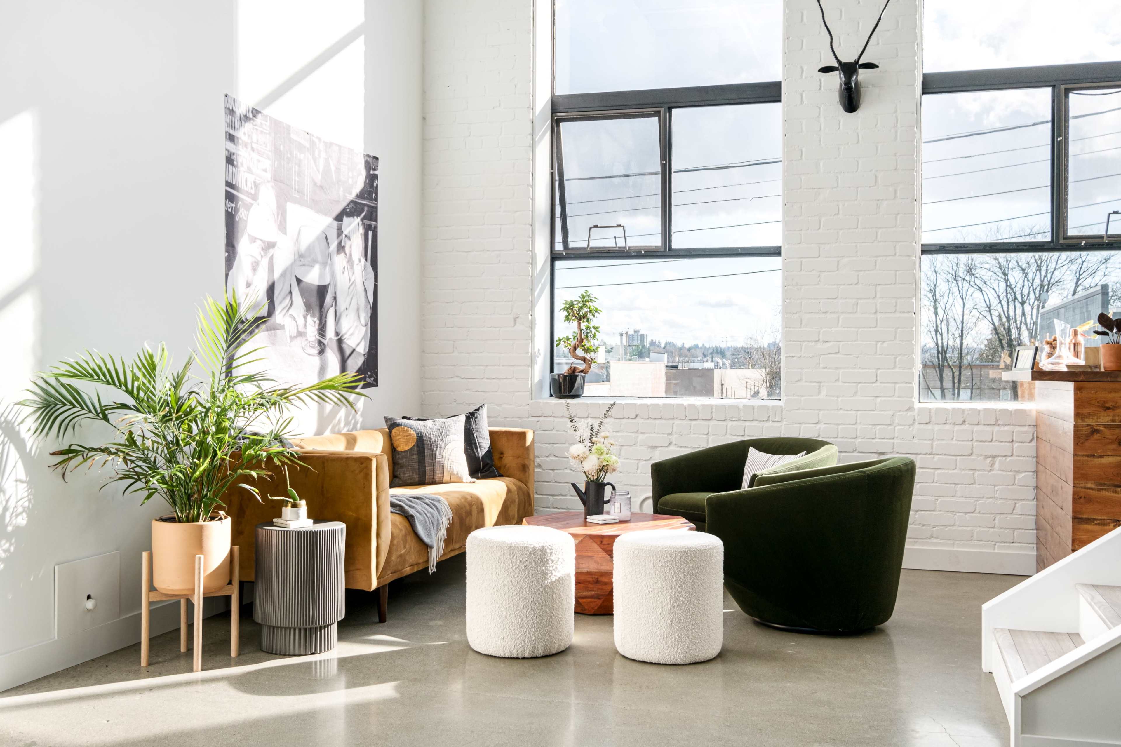 A bright, modern living space featuring a brown couch, green armchair, two white poufs, a wooden coffee table, and a potted plant near large windows.