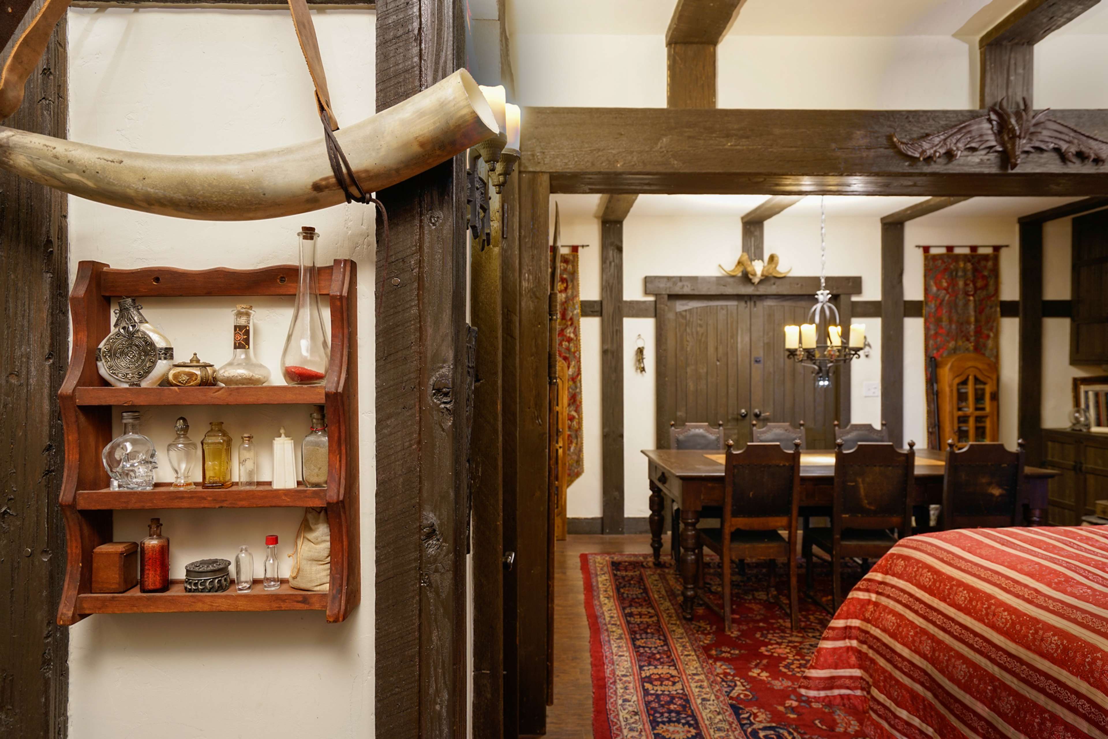 The image shows a rustic interior featuring a wooden shelf with various glass bottles, a dining table with chairs set against a backdrop of decorative wall hangings, and a patterned rug on the floor.