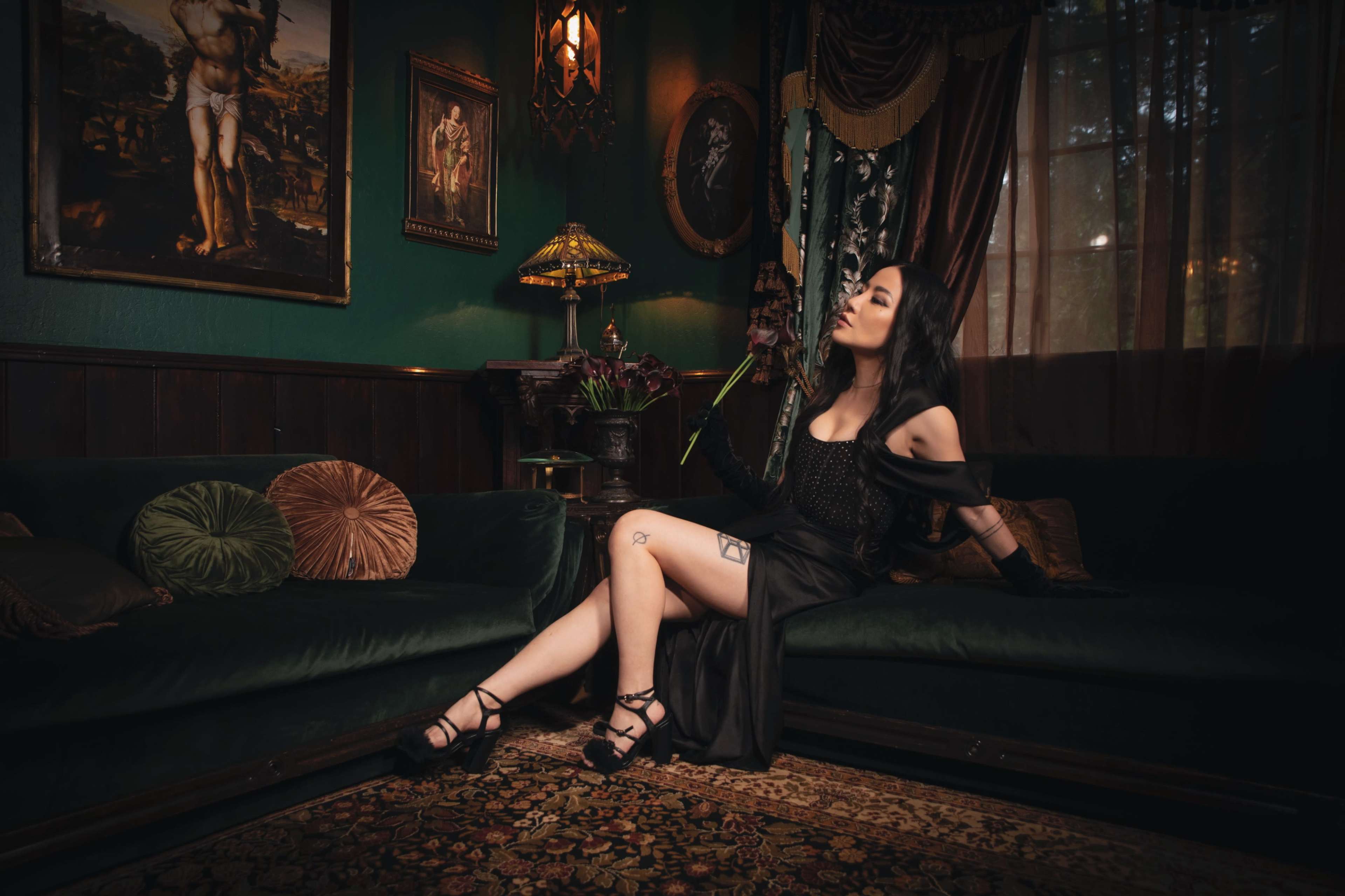 A woman in a dark outfit poses on a green velvet sofa in a richly decorated room with art on the walls and a patterned rug.