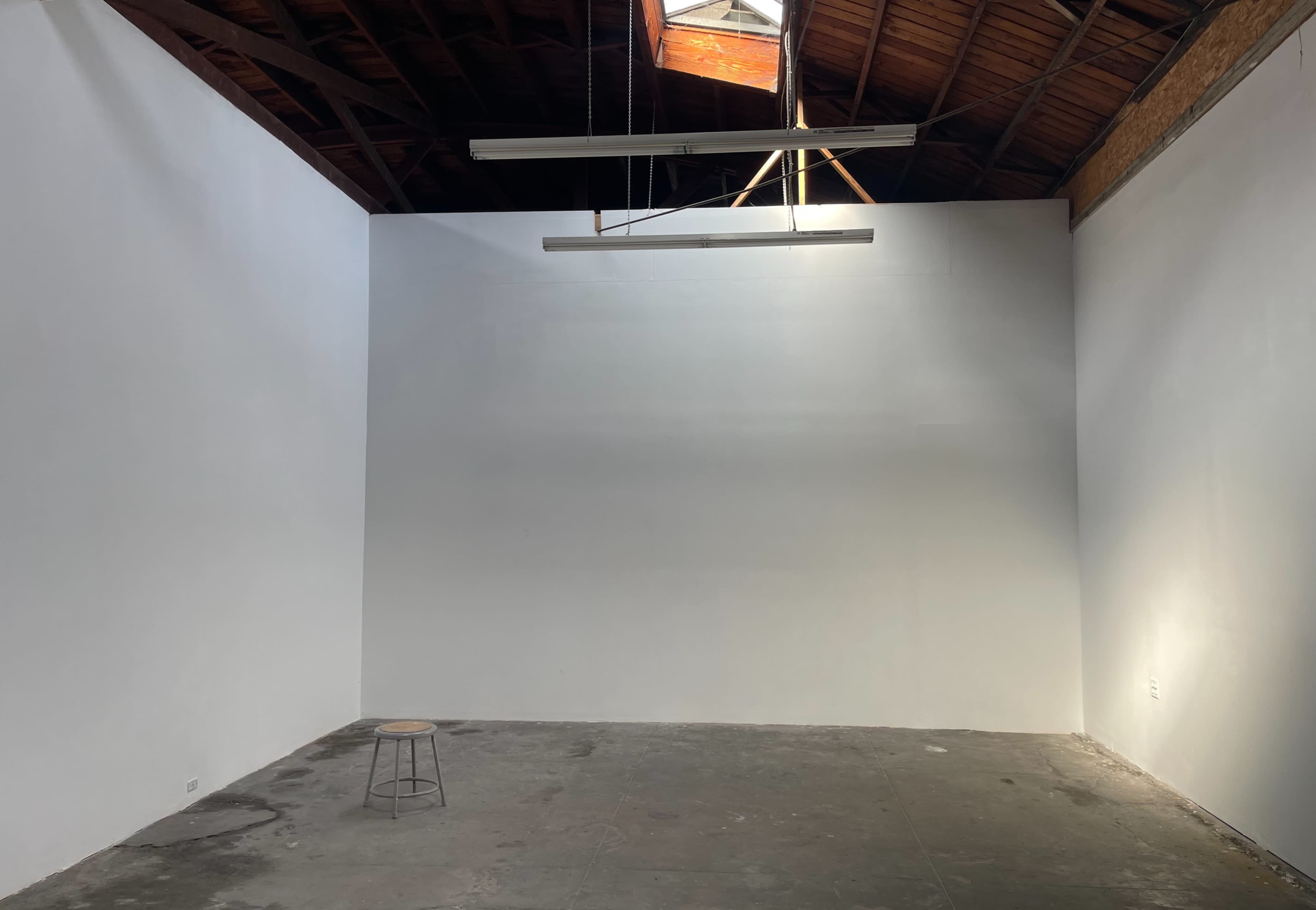A spacious, vacant room with white walls, a concrete floor, a wooden ceiling, and a single stool in the center.