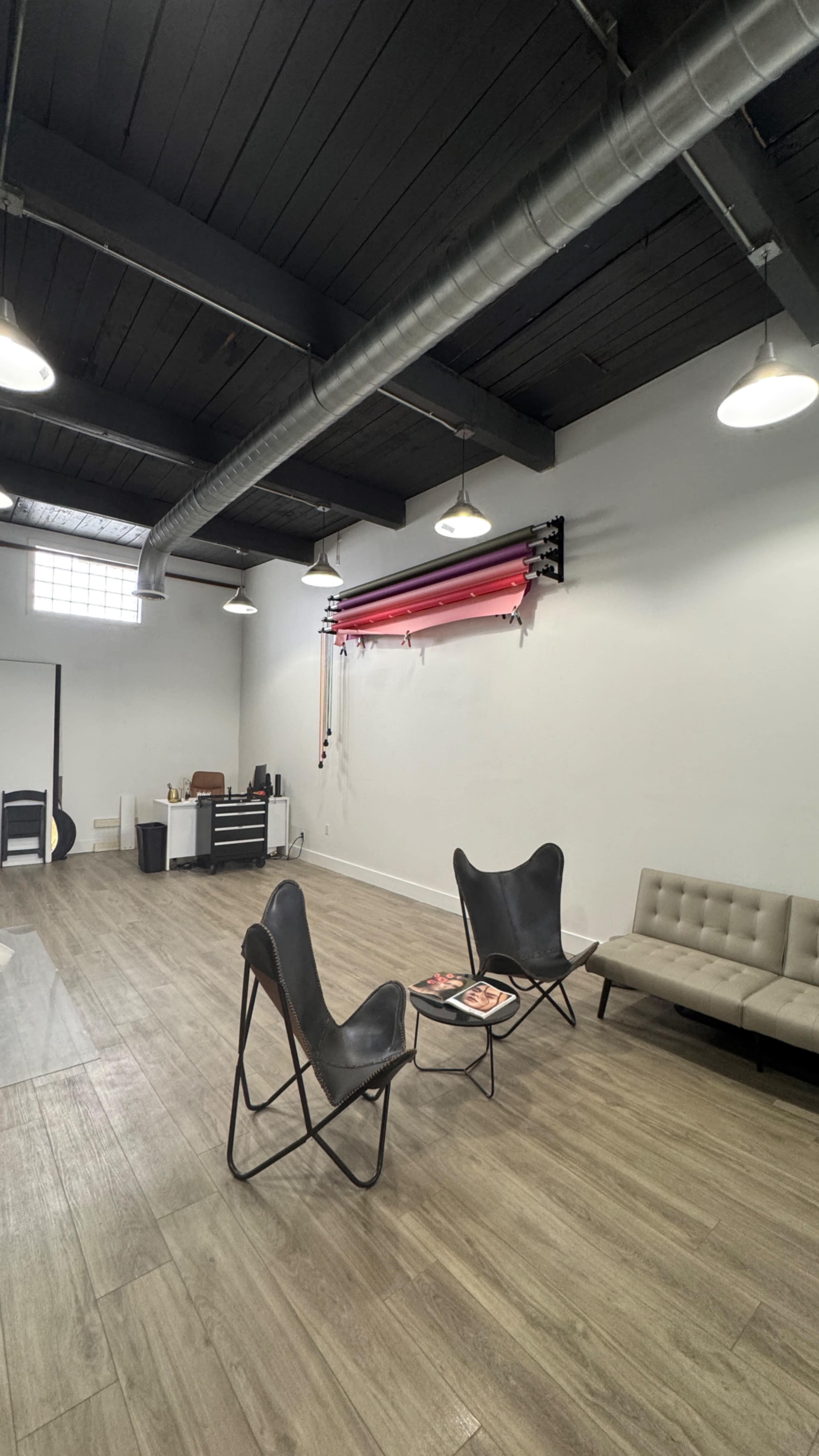 The image shows a spacious studio with gray walls, wooden flooring, and minimalist furniture, including two black chairs and a light-colored sofa.