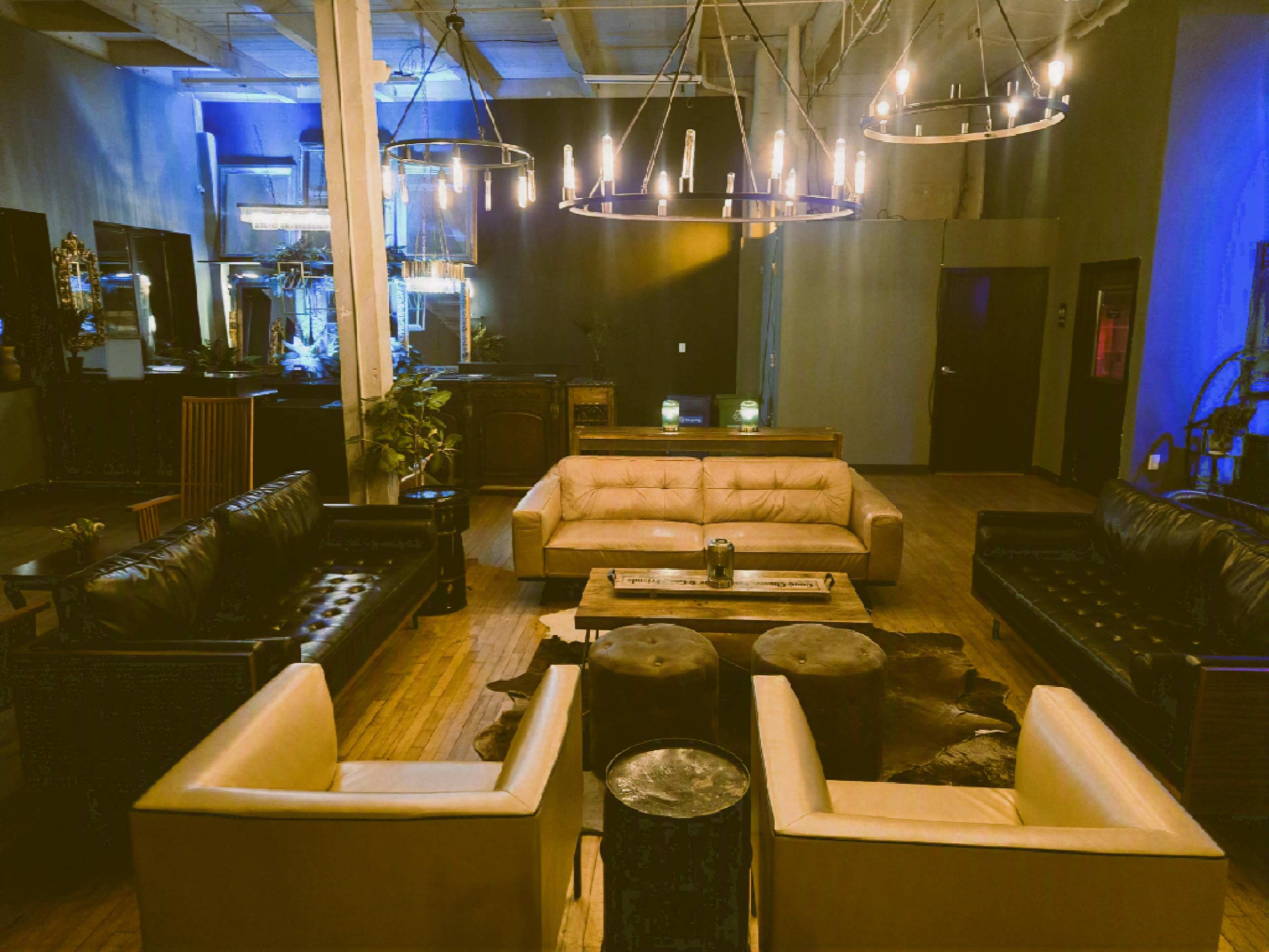 The image shows a spacious lounge area with a combination of leather sofas arranged around a coffee table, illuminated by modern chandelier lighting against a dimly lit backdrop.