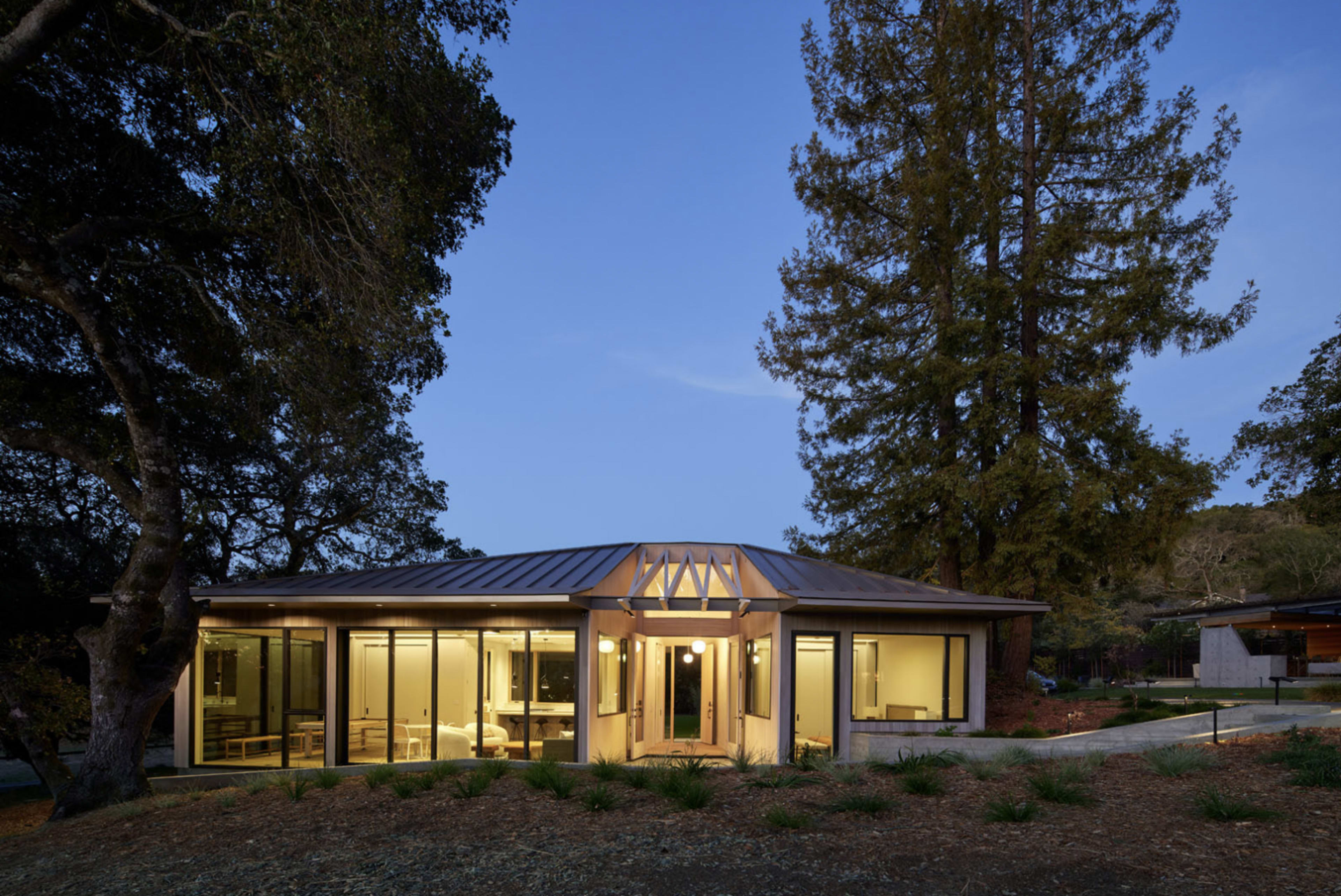 A modern, single-story house with a metal roof features large glass windows, surrounded by trees and landscaped grounds at dusk.
