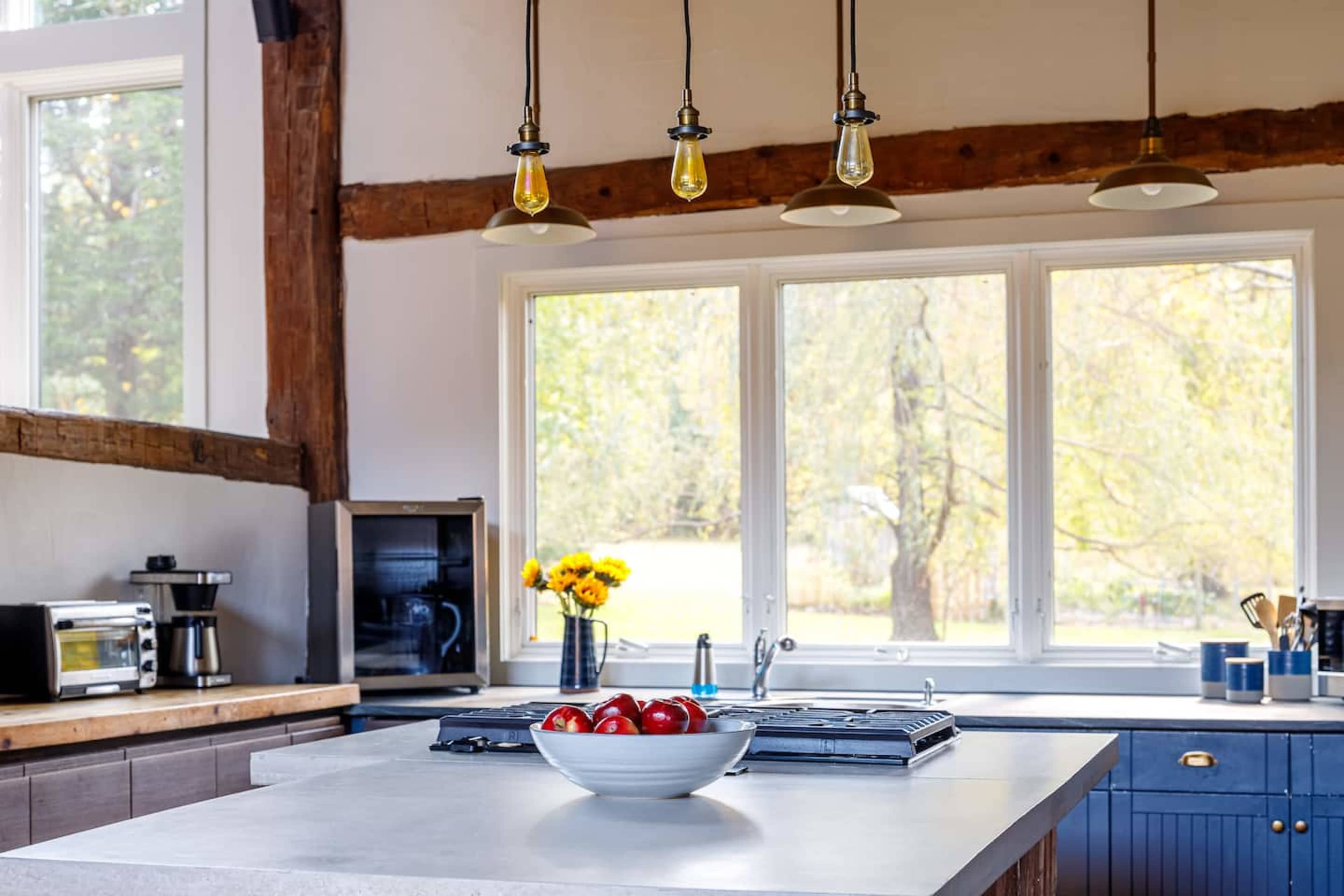 A modern kitchen features a central island with a bowl of red apples, large windows, and hanging light fixtures.