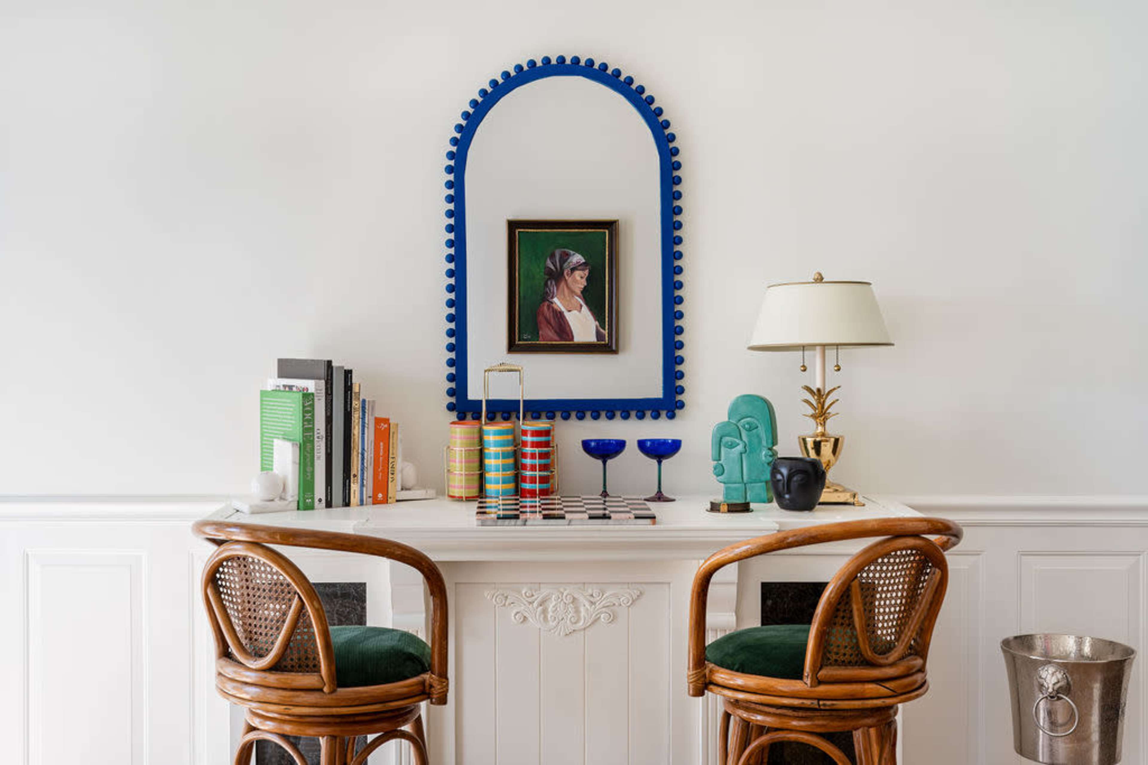 A console table with two rattan chairs displays a framed portrait, books, decorative items, and a lamp against a white wall.