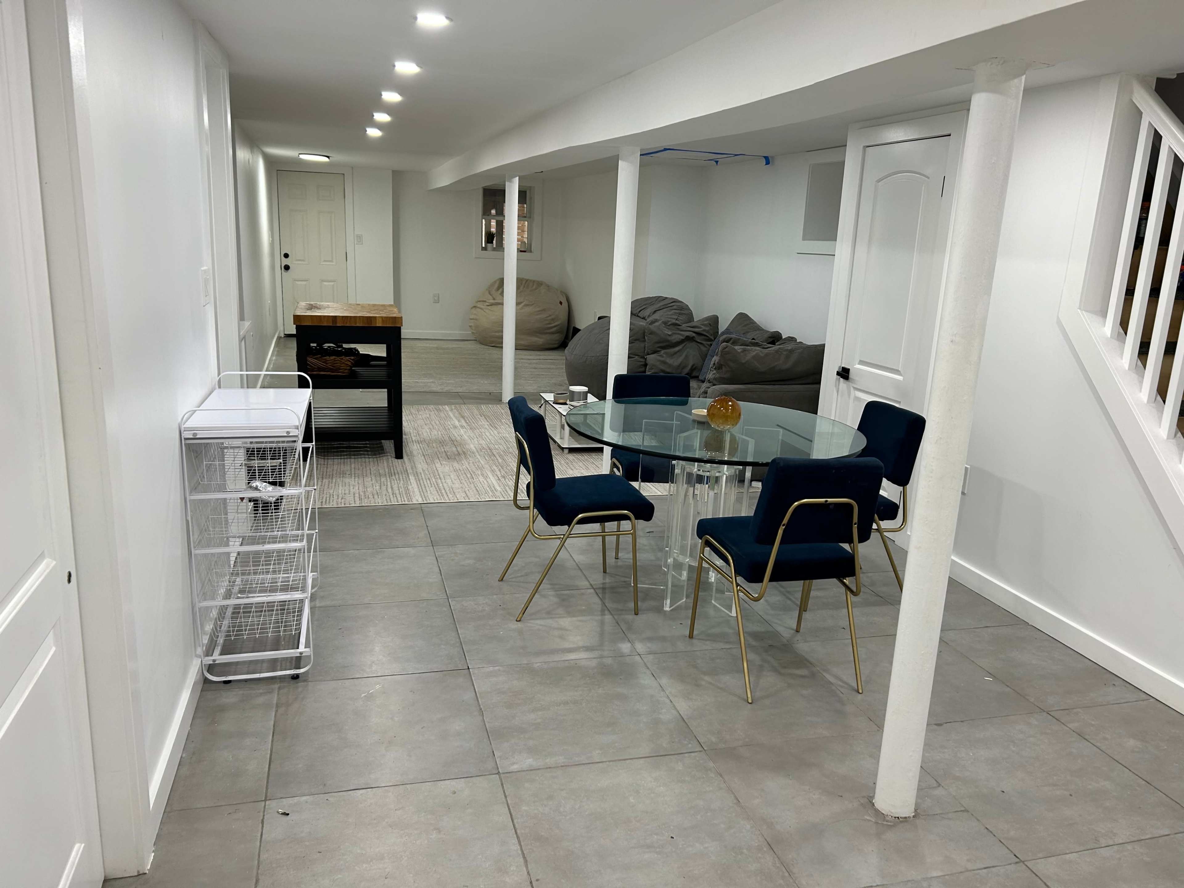 The image shows a modern basement space featuring a glass dining table surrounded by four blue chairs, a laundry cart, and bean bags in the background.
