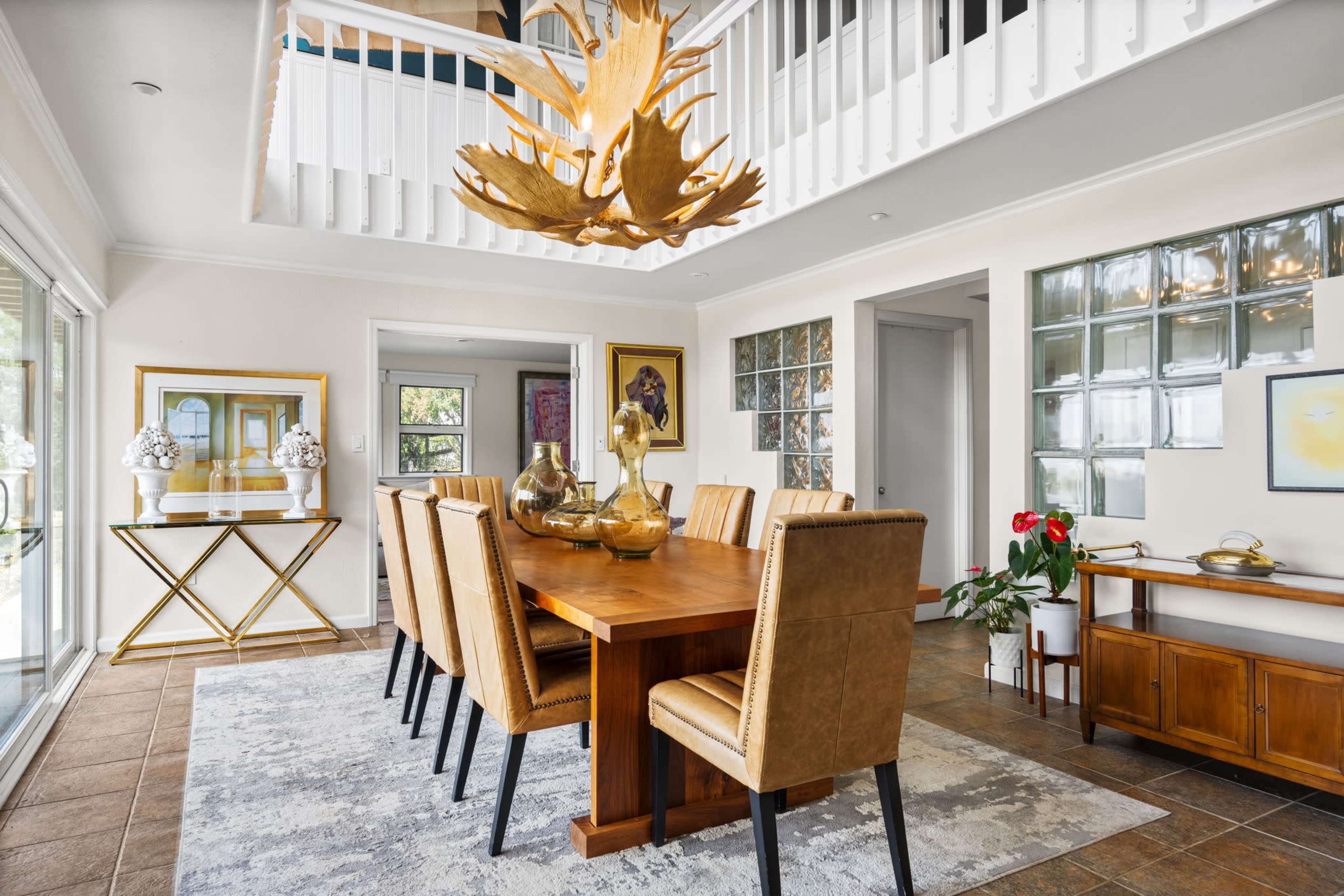 A bright dining room features a wooden table surrounded by upholstered chairs, with a large chandelier and artwork adorning the walls.