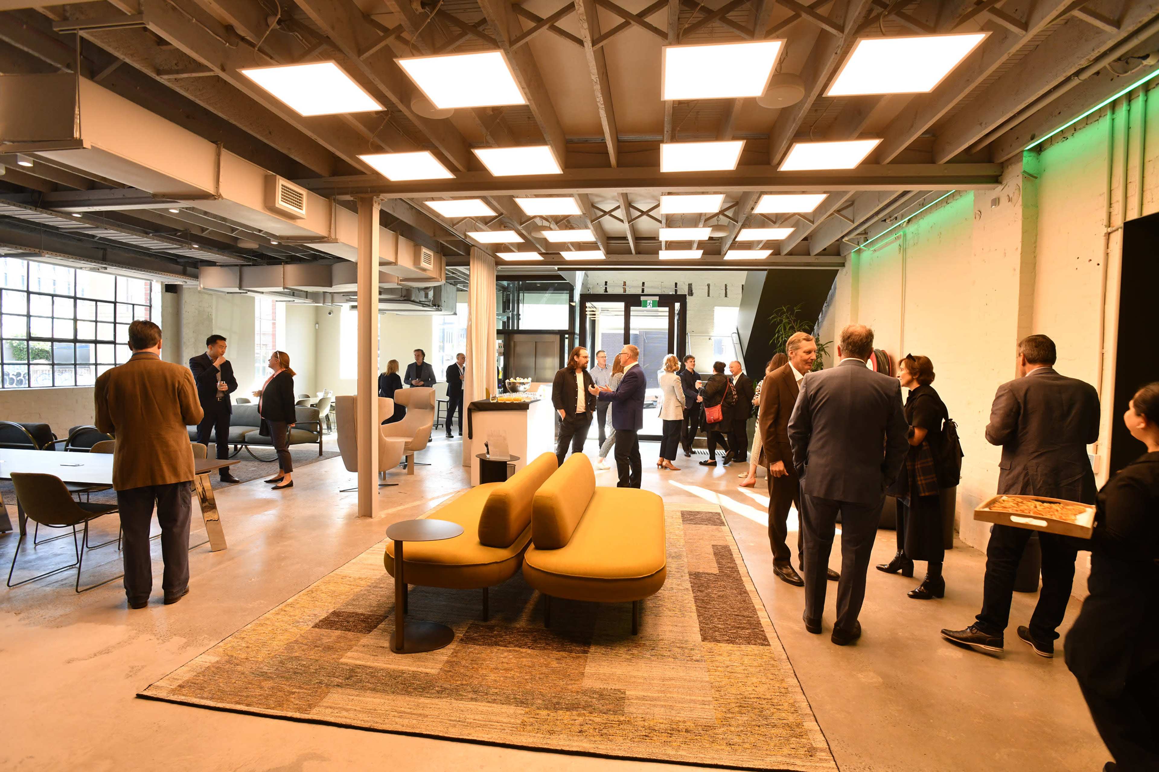 The image shows a modern office space with people mingling, a large area rug, and contemporary furniture arranged for a gathering.