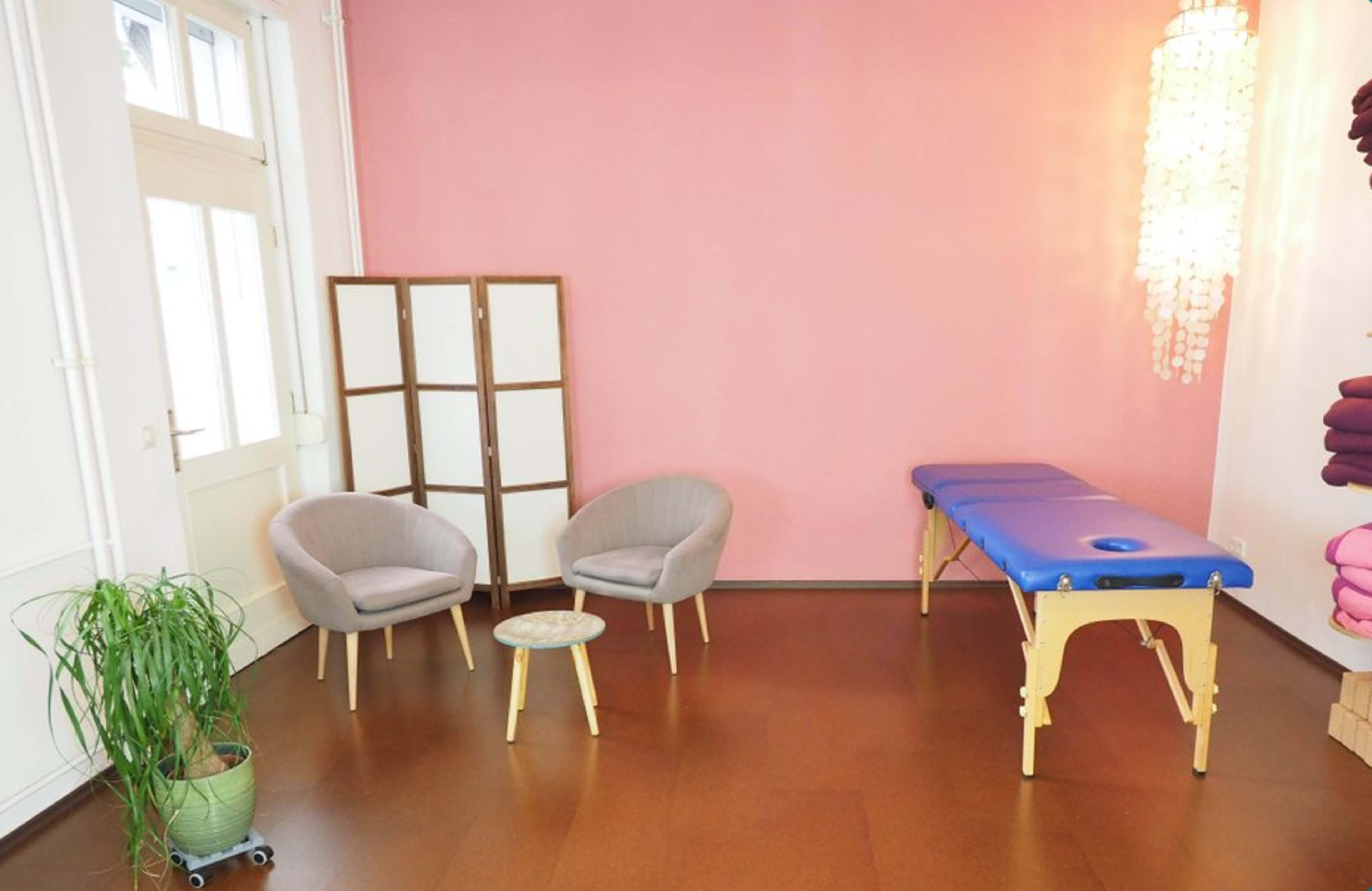 A massage therapy room features two gray chairs, a small table, a treatment table with a blue cover, and a pink wall.