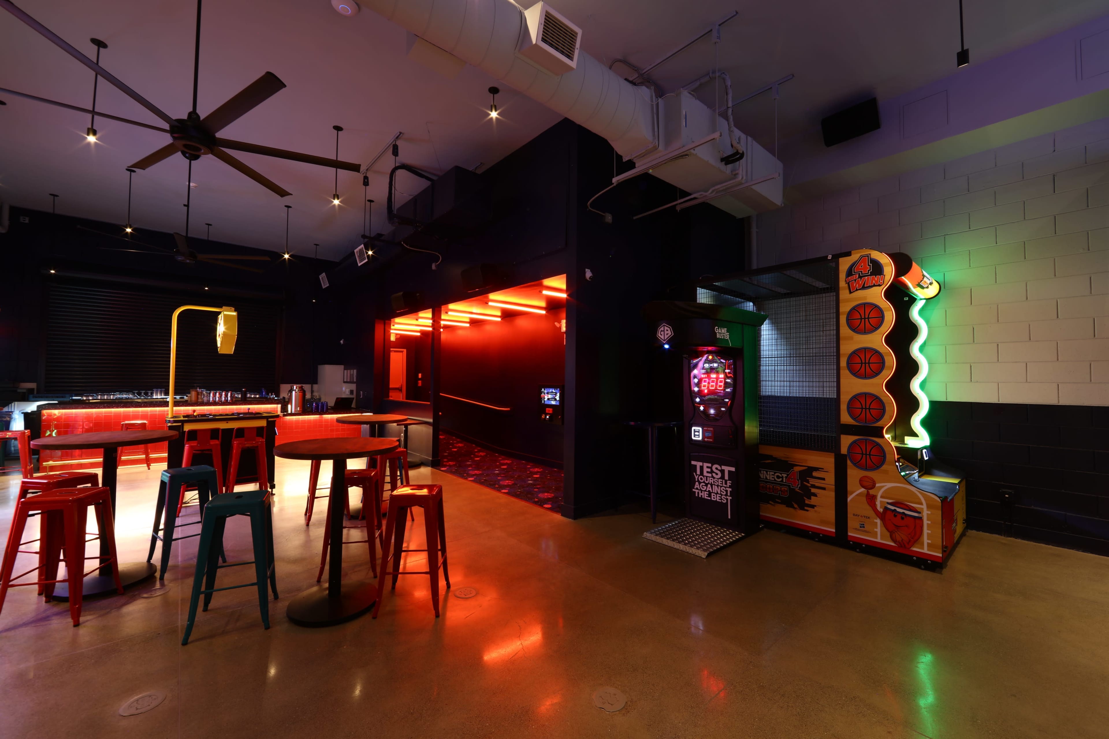 The image shows a modern bar area with colorful stools, a game machine, and bright neon lights accentuating the space.