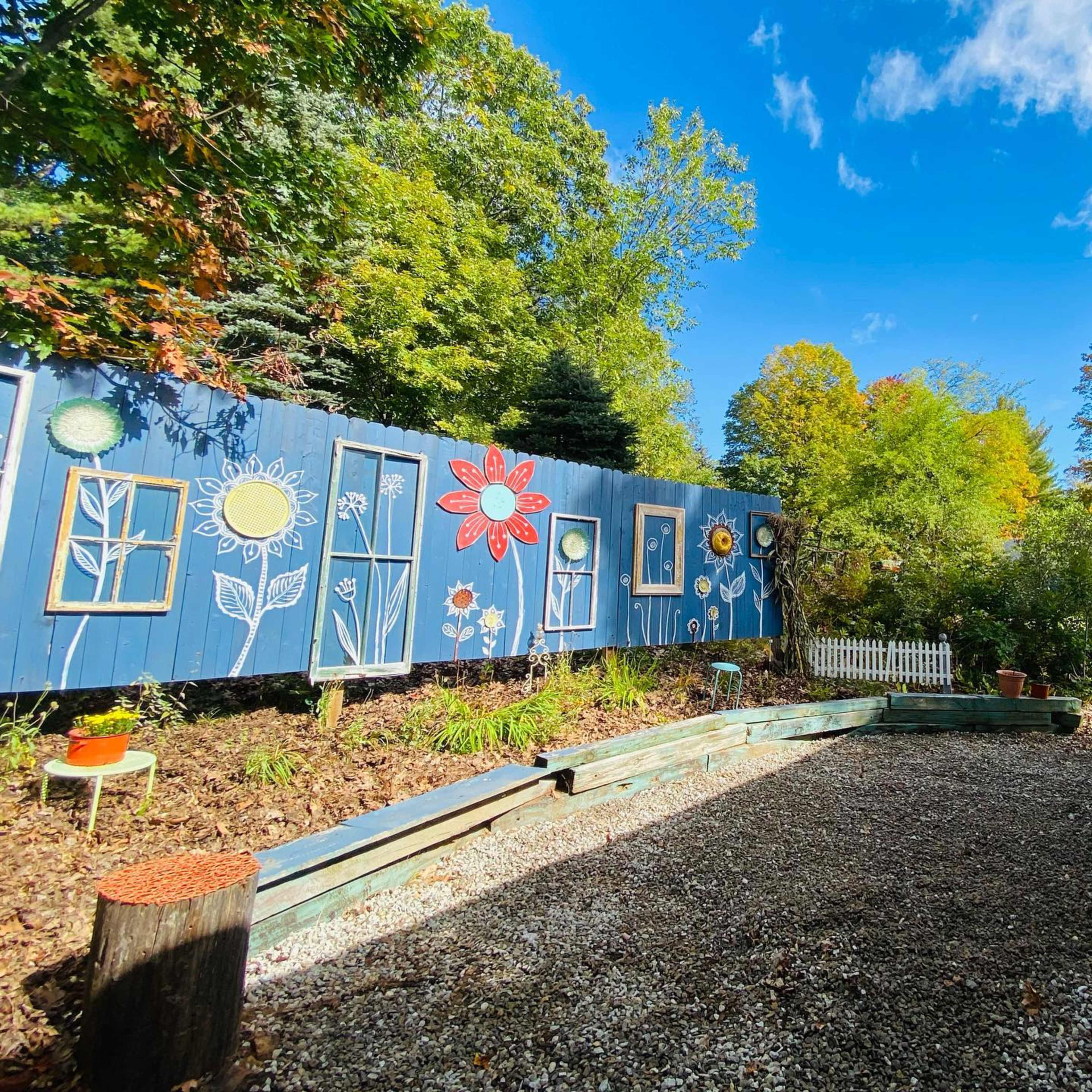 A colorful mural of flowers and framed windows adorns a wooden fence in a garden setting with gravel paths and greenery.