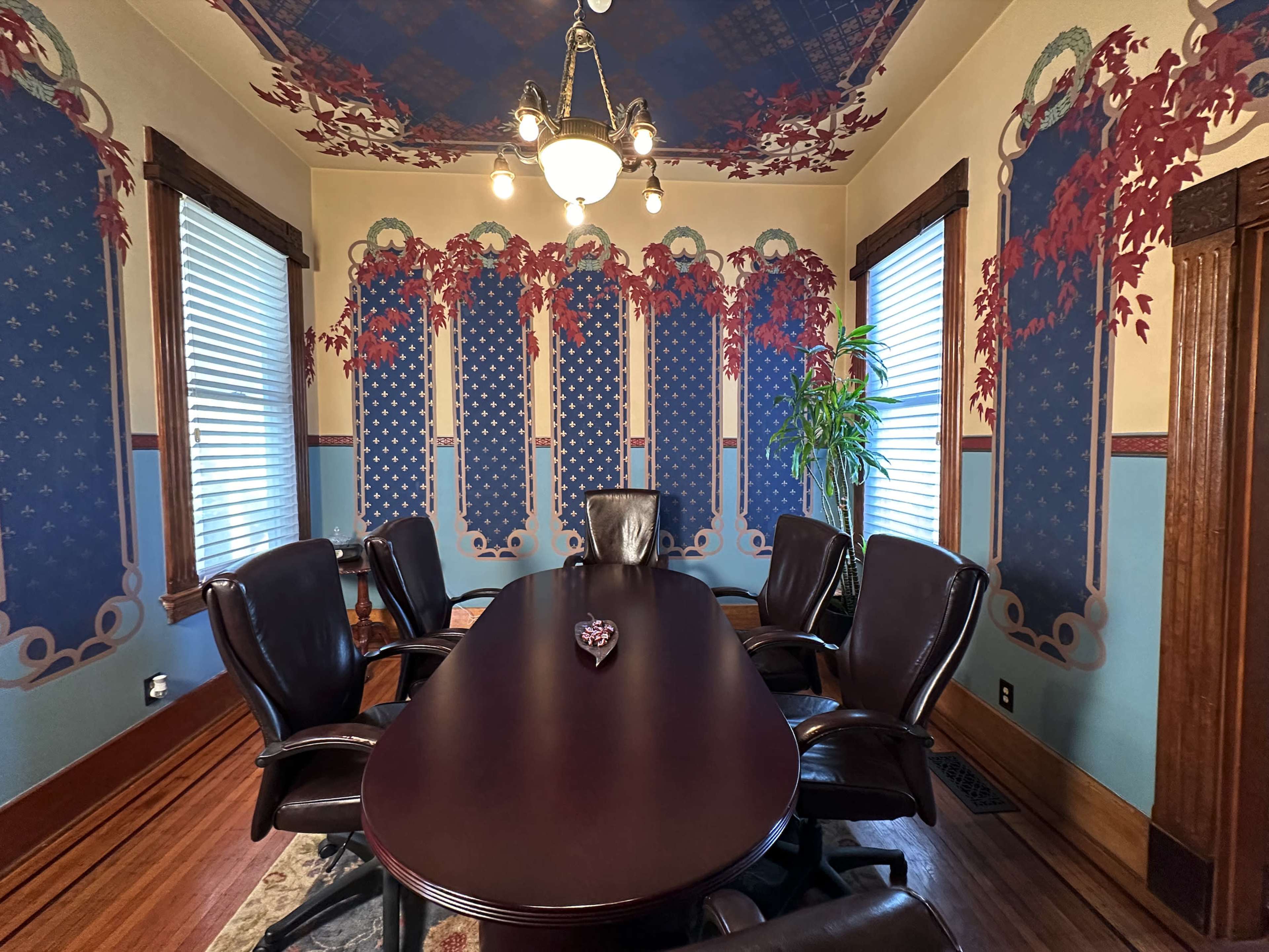 A conference room features a large oval table surrounded by brown leather chairs, with intricately designed wallpaper depicting floral and geometric patterns.