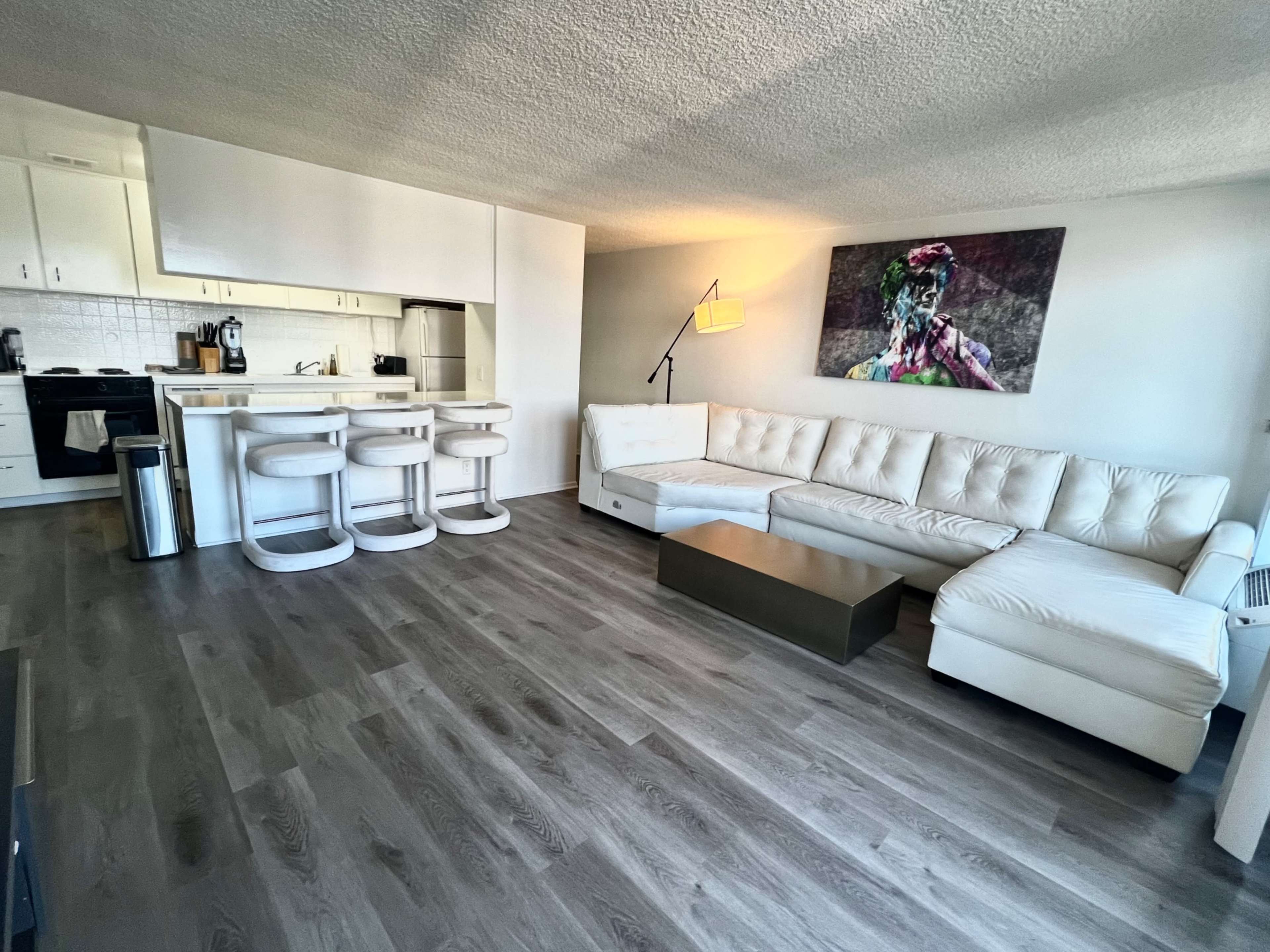 A modern living room features a white sectional sofa, a glass coffee table, and a kitchen bar with three stools.