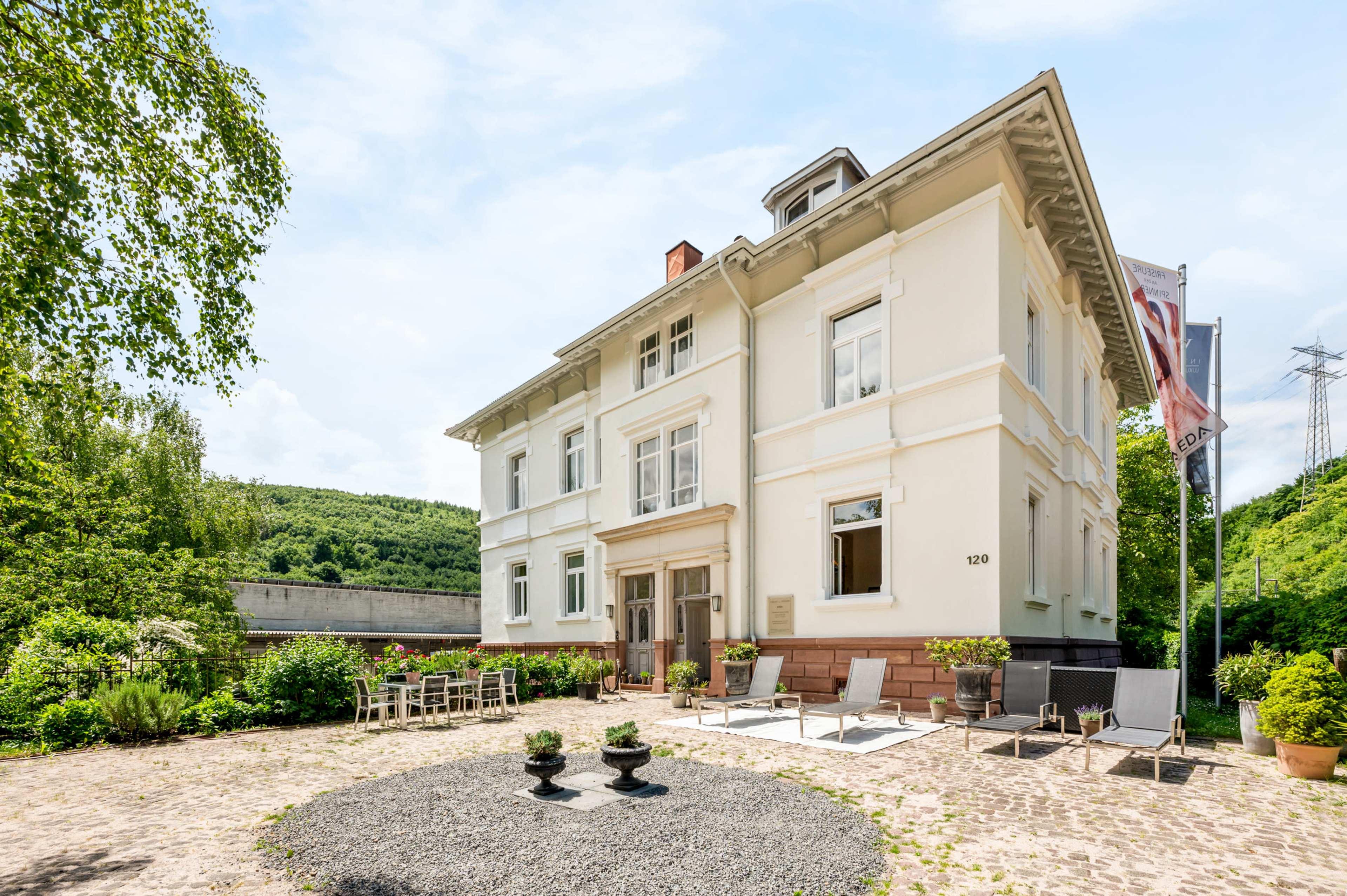 A large, two-story white building with a decorative facade is set in a garden area featuring outdoor seating and potted plants.