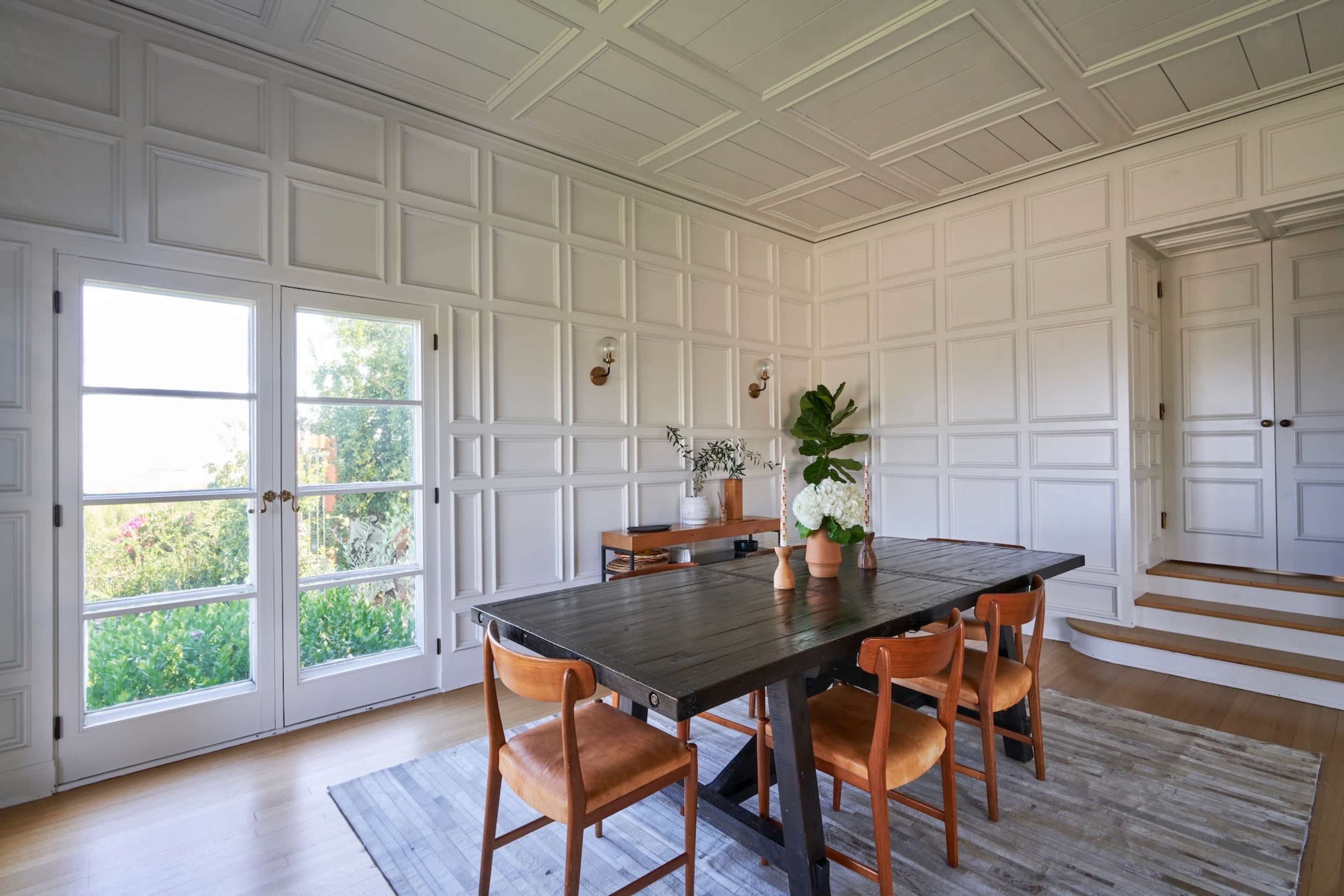 A dining room features a wooden table surrounded by chairs, with a set of French doors opening to a garden view and paneled walls.
