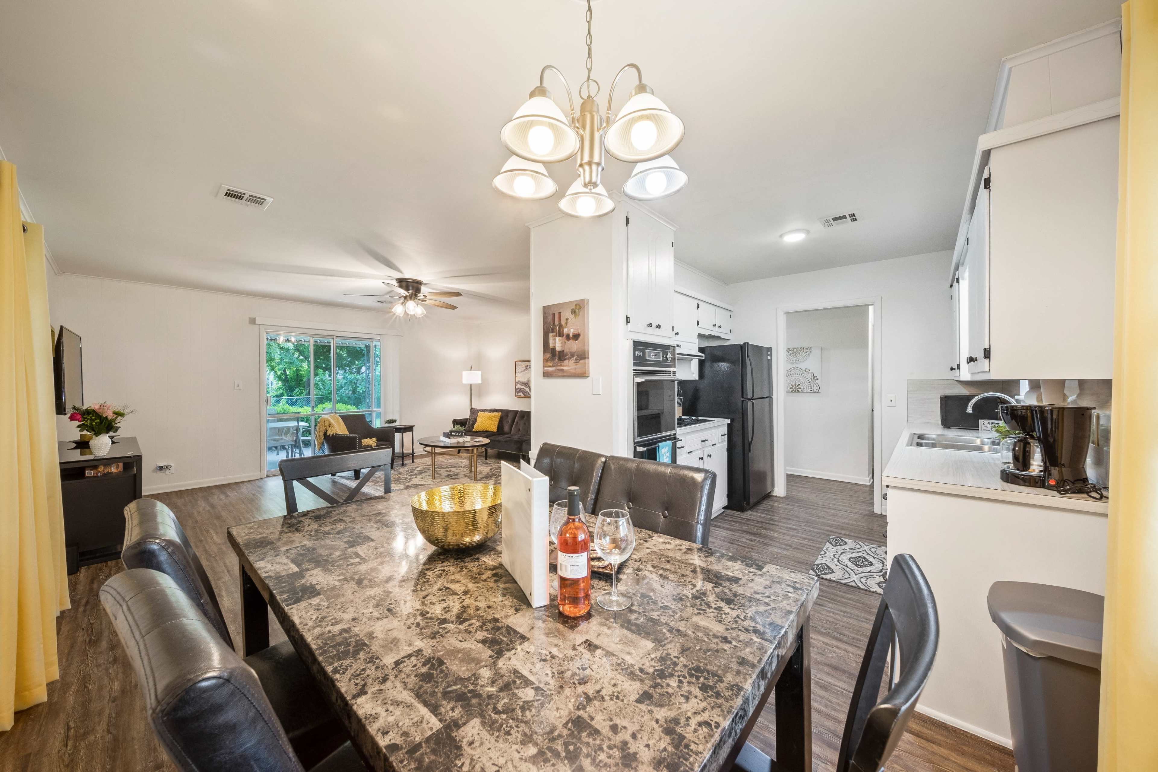 A modern kitchen and dining area featuring a dark marble table with eight chairs, a chandelier overhead, and a living space visible in the background.