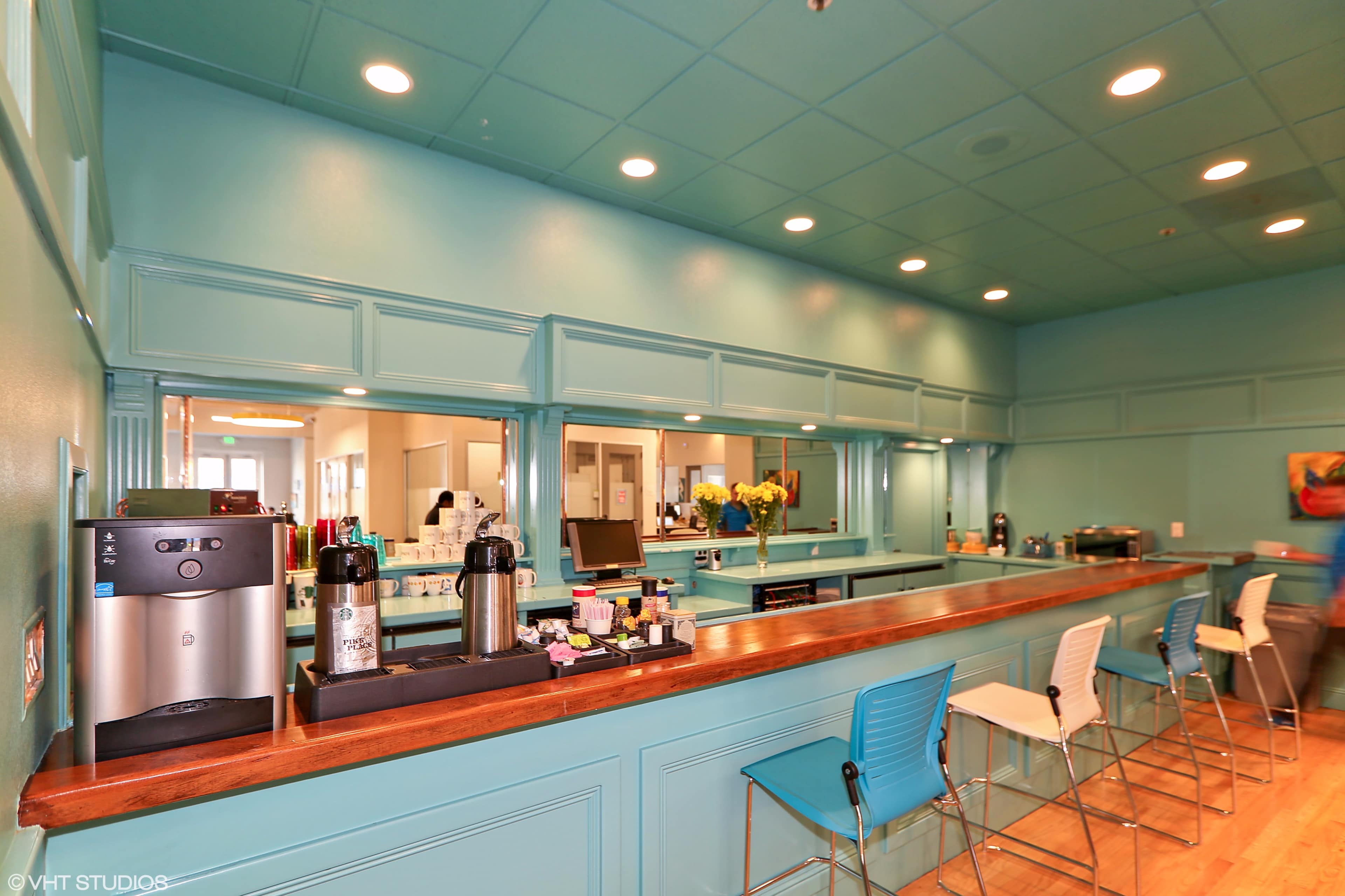 A brightly colored room with a turquoise wall, a wooden counter, and several coffee machines and seating.