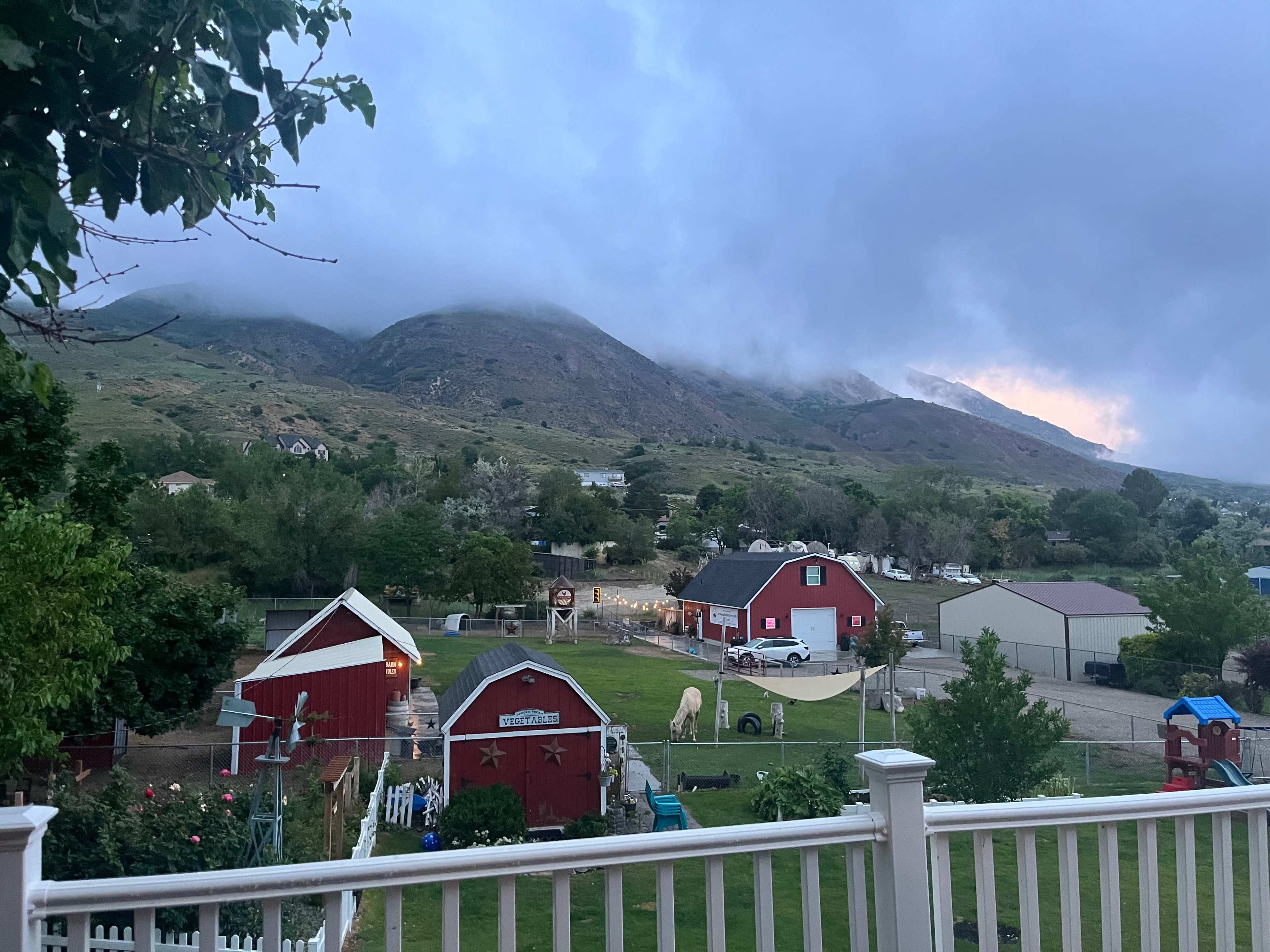A rural scene with a view of mountains in the background, a cloudy sky, and a yard with a barn and several houses situated nearby.