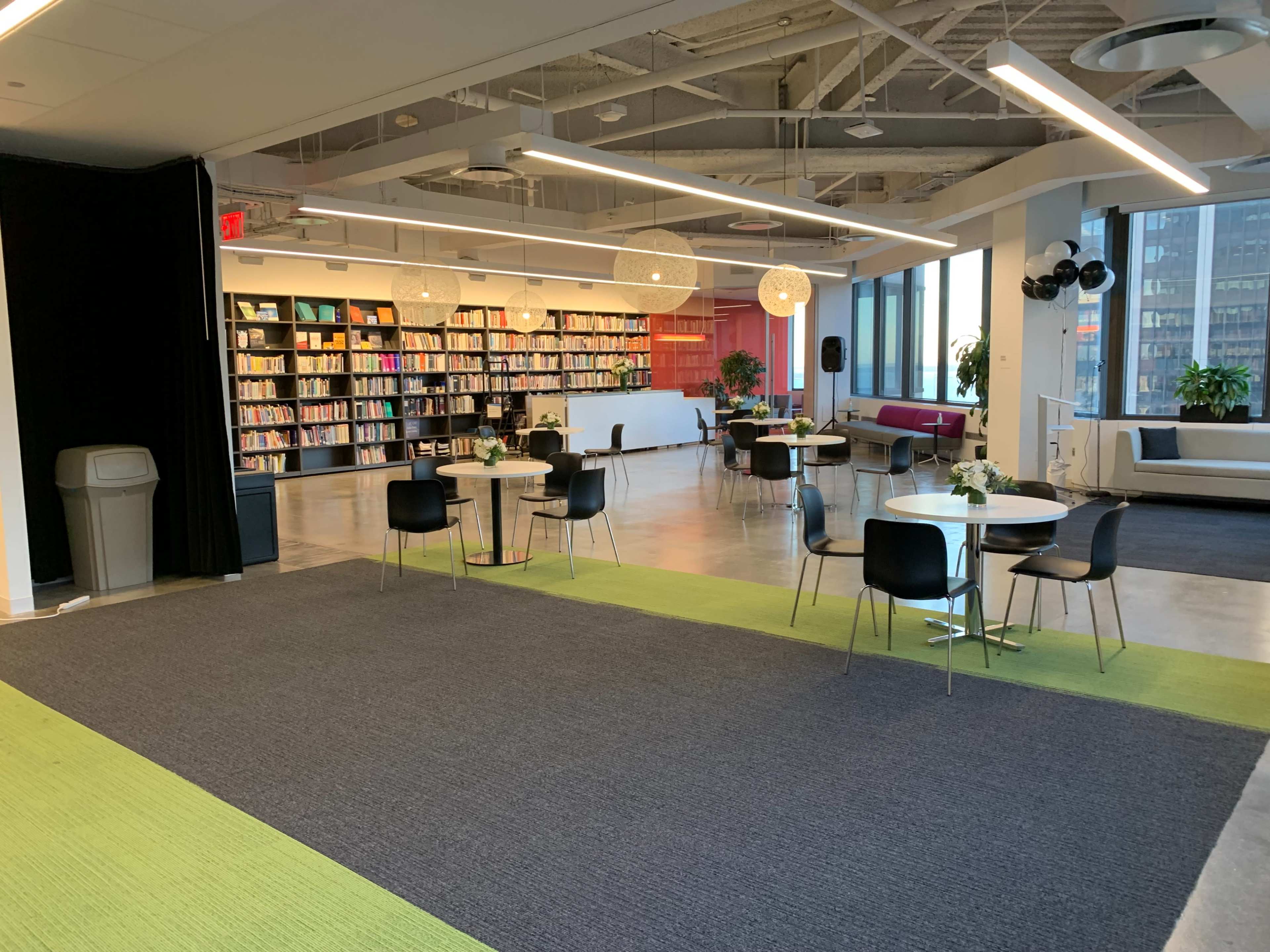 A spacious, modern room with tables and chairs arranged around a carpeted area, a wall of bookshelves in the background, and large windows allowing natural light.