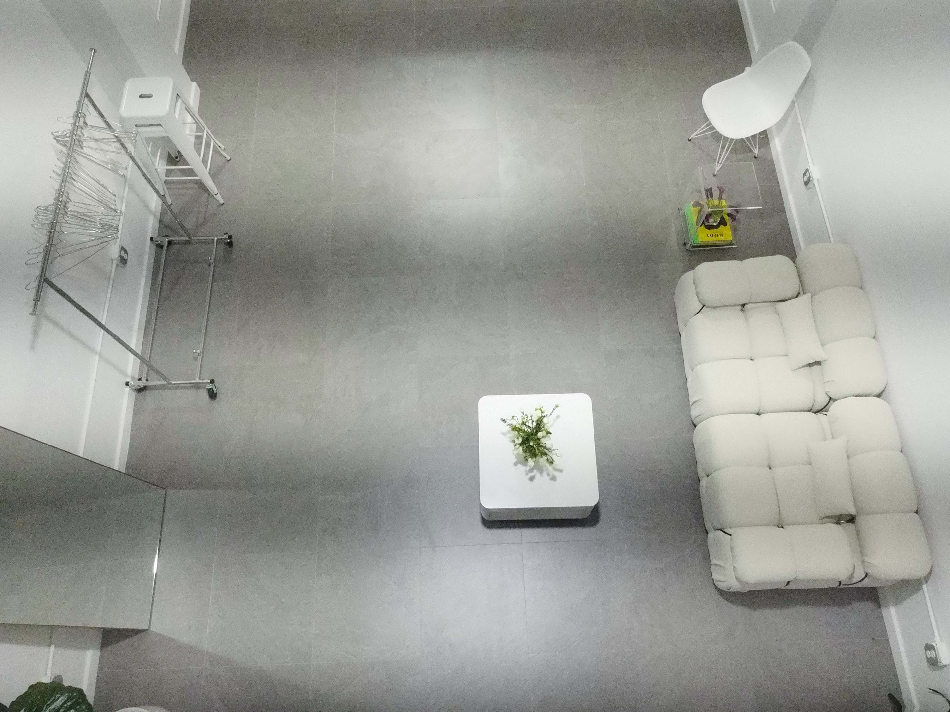 The image shows a minimalistic room with a light-colored couch, a small white coffee table with a plant, a clothes drying rack, and a bar stool in a corner.