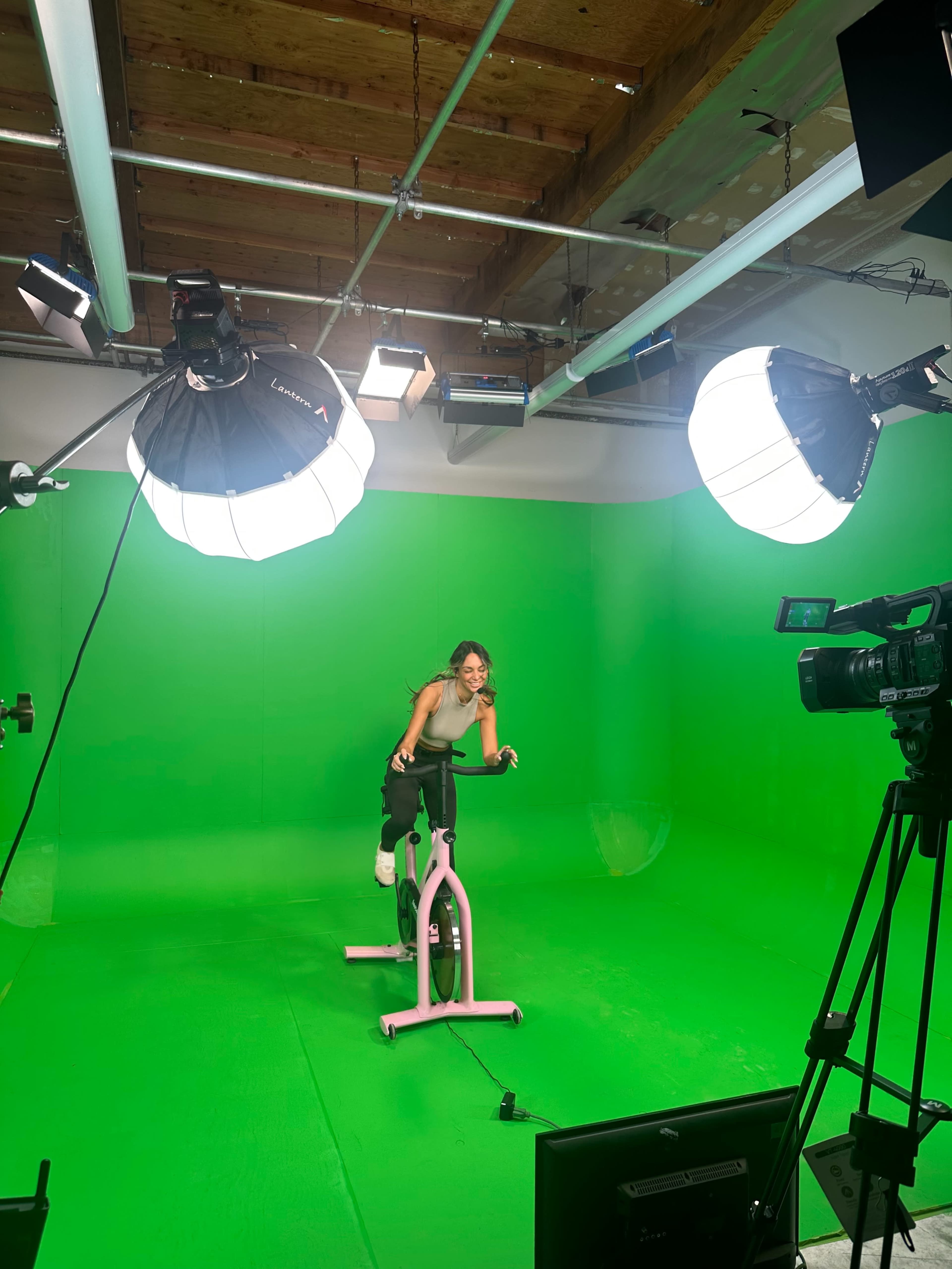 A woman is posing on a stationary bike in a studio equipped with a green screen and professional lighting.