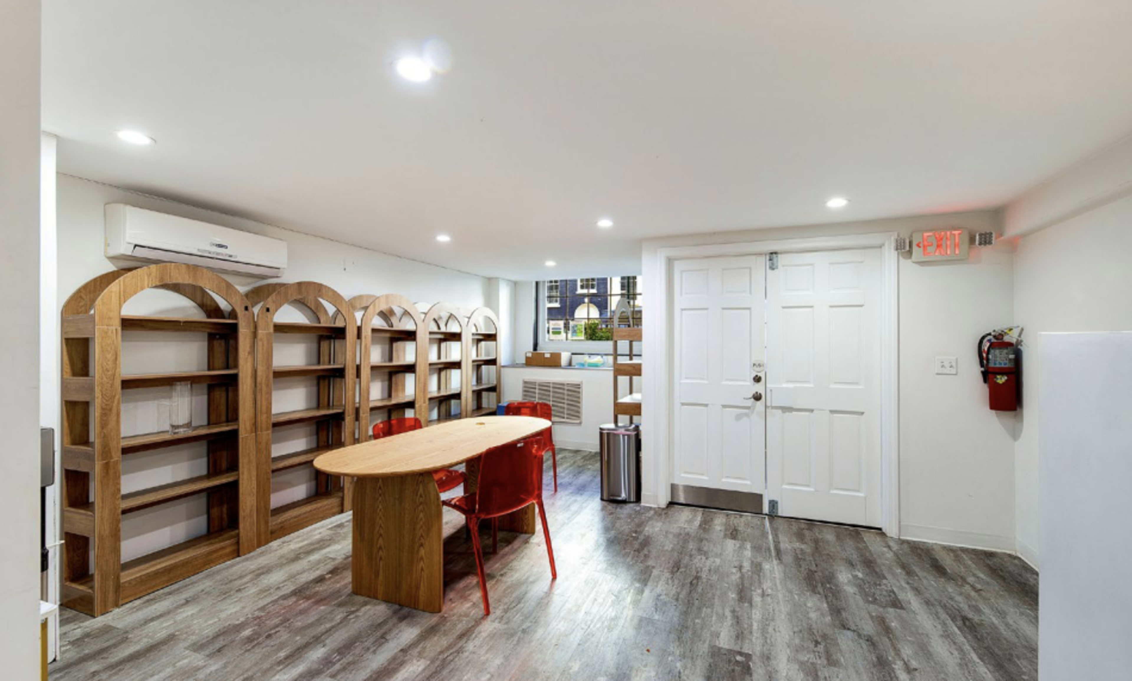 The image shows a tidy room with wooden bookshelves, a round table, and red chairs, illuminated by overhead lights.