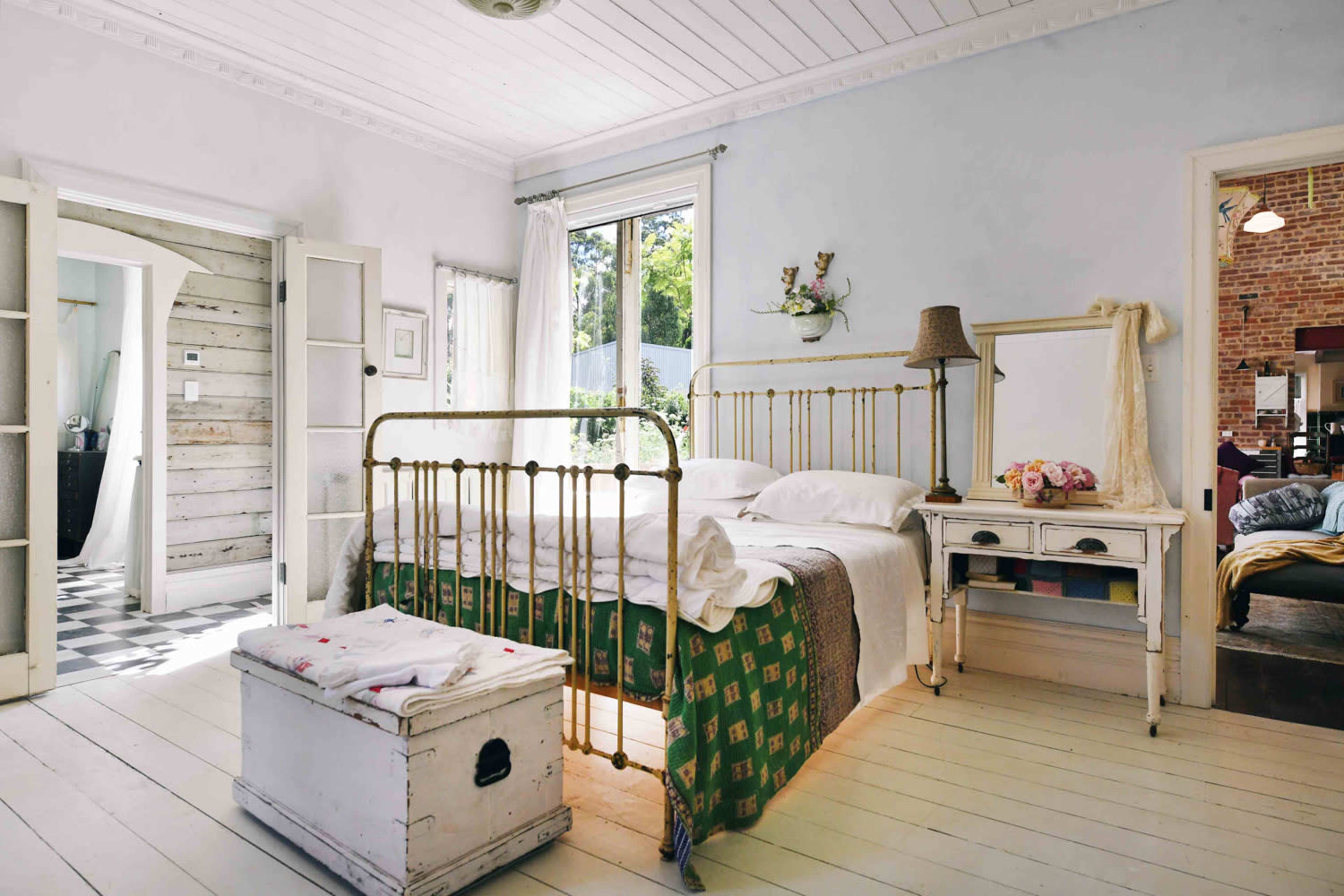 A softly lit bedroom with a brass bed frame, a green patterned blanket, and white wooden flooring, complemented by a vintage storage chest and a window overlooking greenery.
