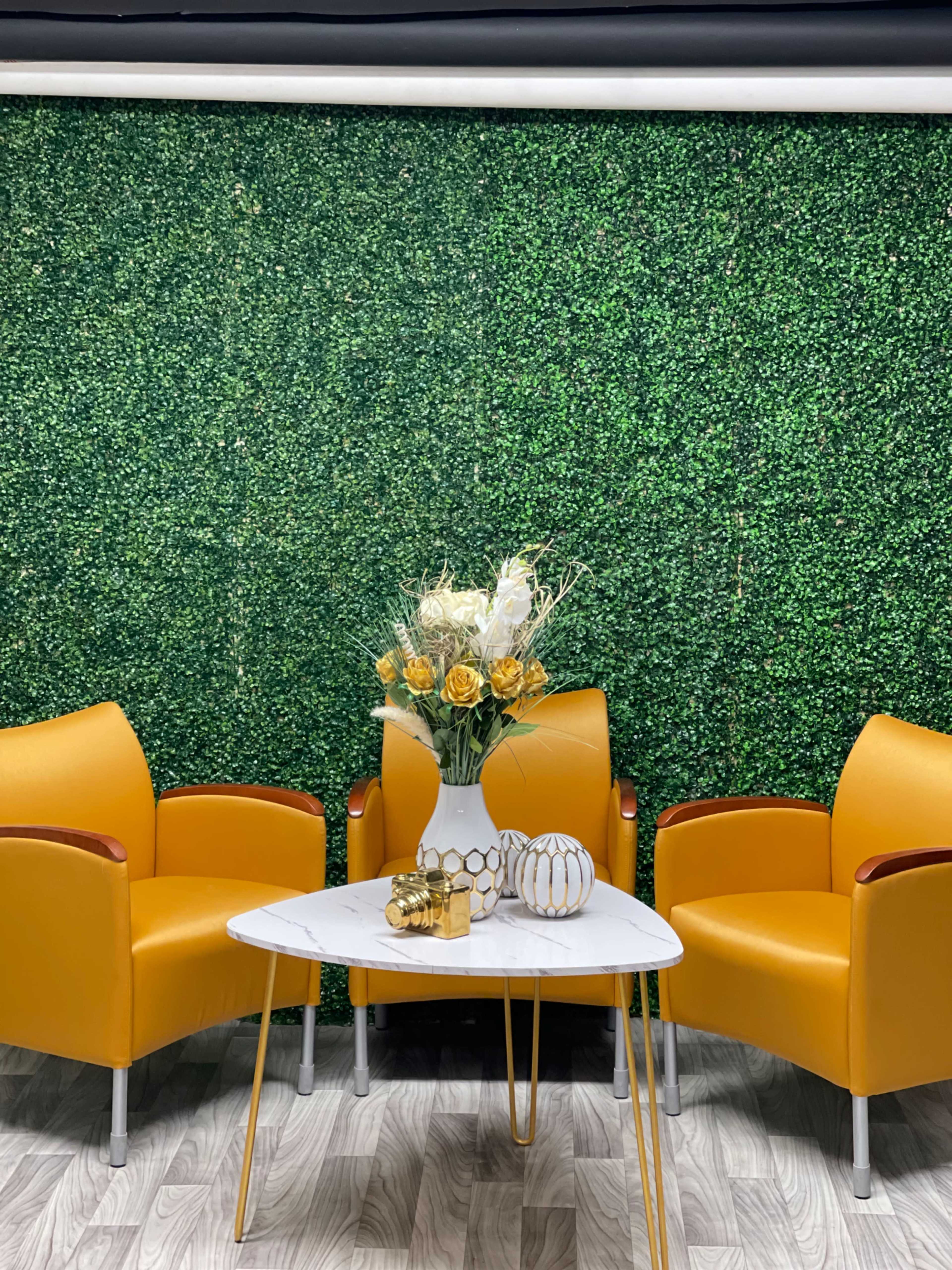The scene features two yellow armchairs flanking a white marble coffee table topped with a floral arrangement, set against a green hedge backdrop.