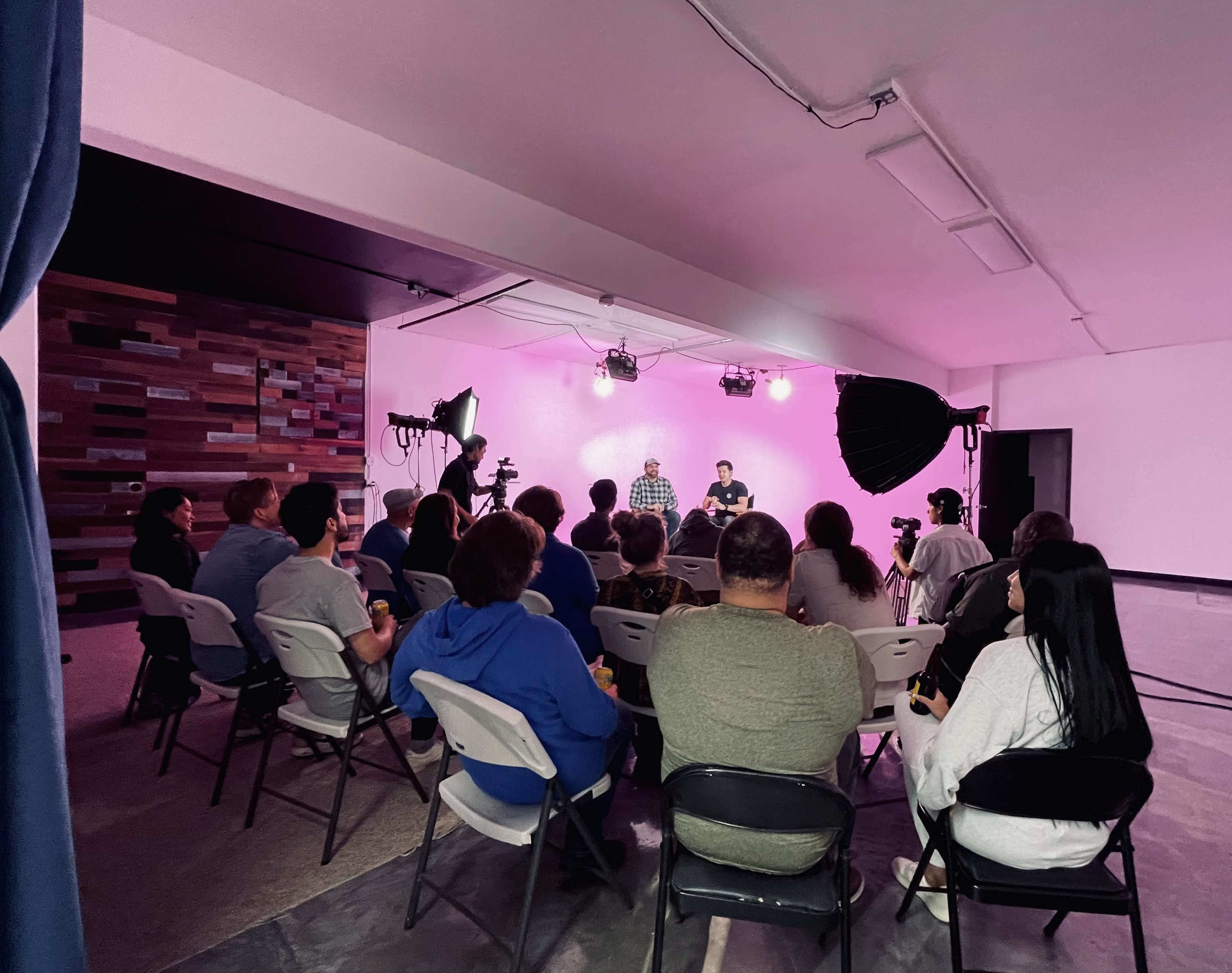 A group of people sit on folding chairs in a dimly lit room with a pink backdrop, watching two individuals seated at a table being filmed.