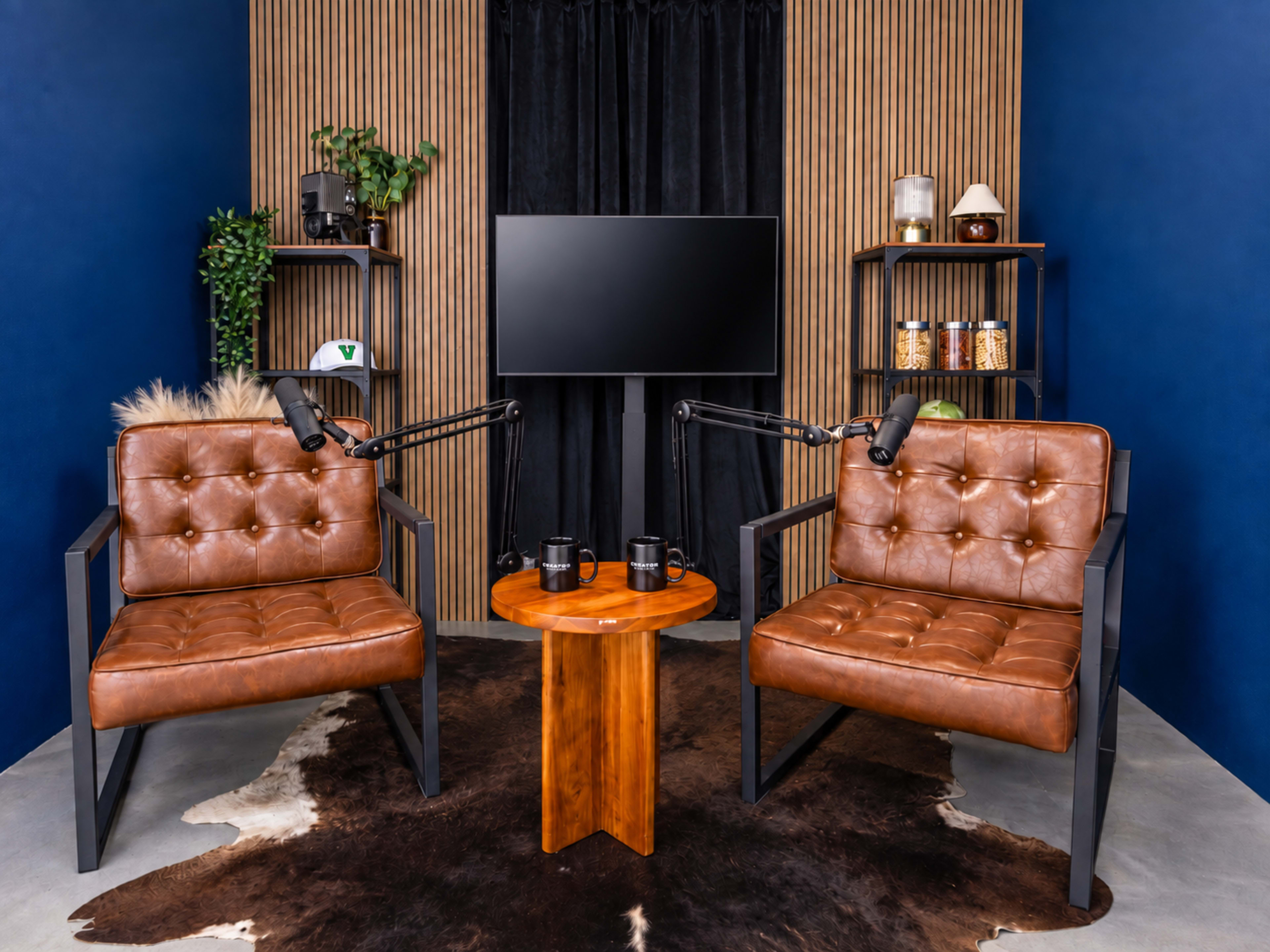 The image features a modern podcast studio setup with two brown leather chairs, a wooden table, and a television screen surrounded by shelves displaying various items.