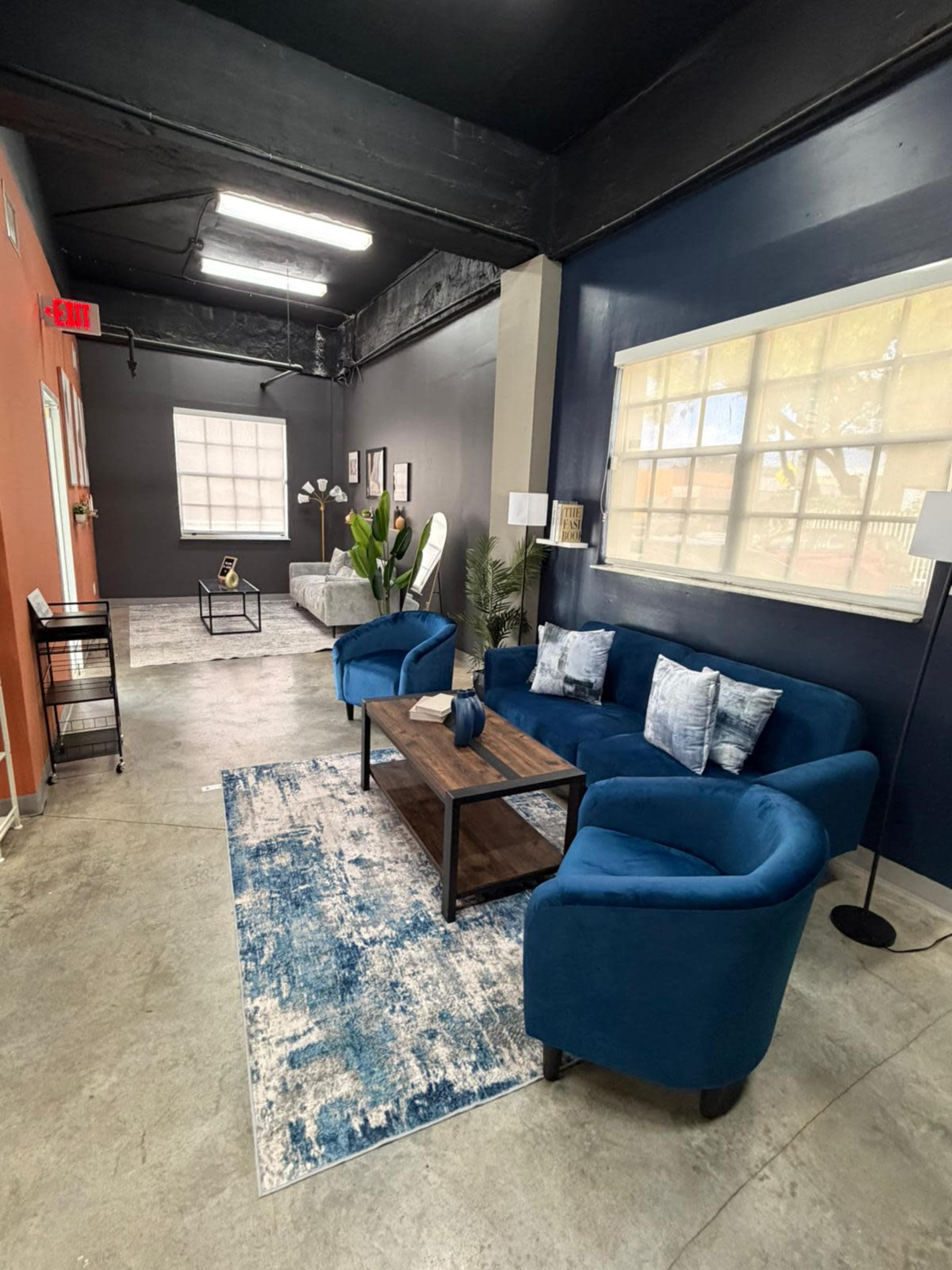 The image shows a modern room with dark walls, a blue seating area consisting of two chairs and a couch, a wooden coffee table, and a decorative rug.