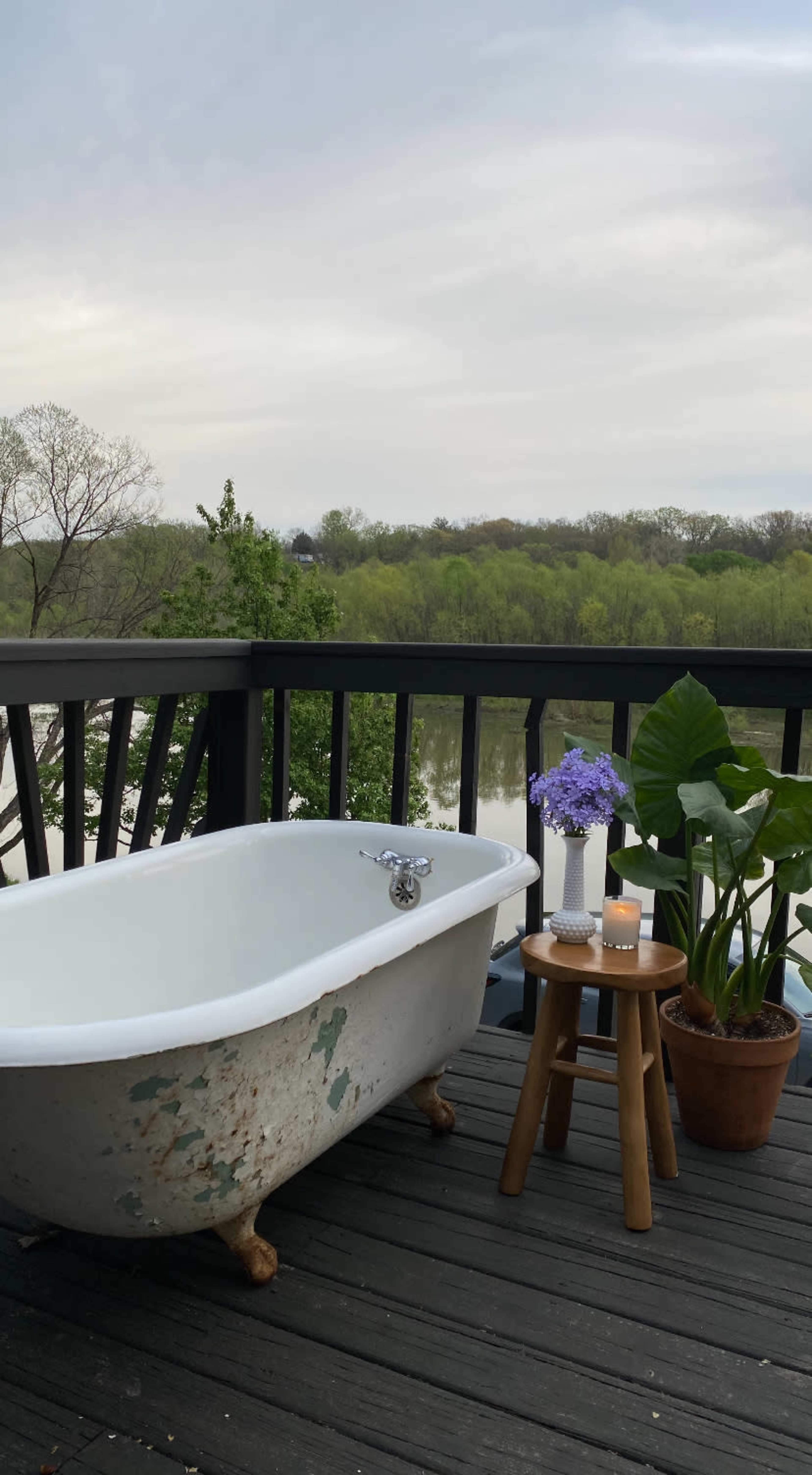 A vintage bathtub sits on a wooden deck overlooking a body of water, accompanied by a small table with a vase of flowers and a potted plant.
