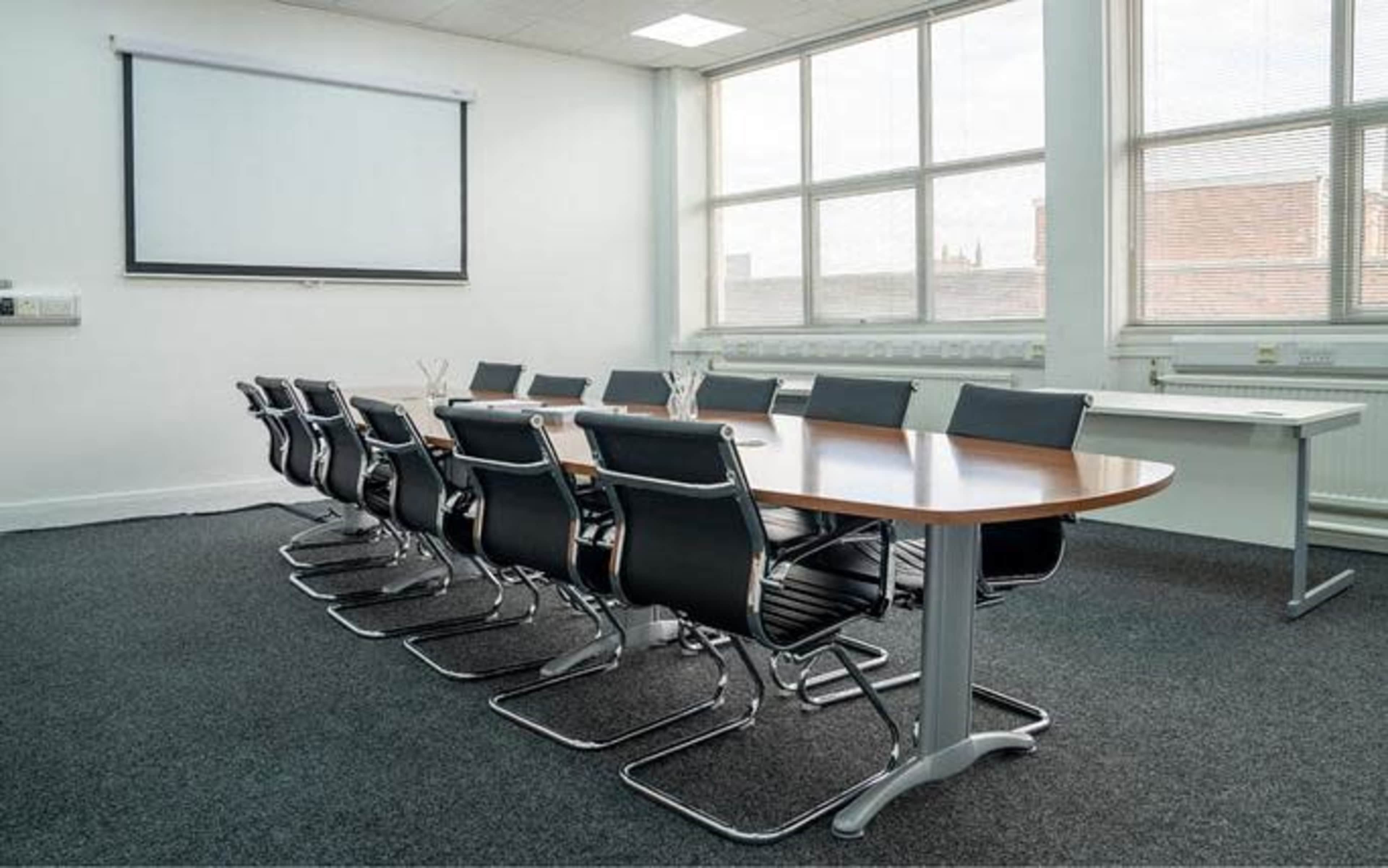 A conference room with a long wooden table surrounded by black ergonomic chairs, featuring a projector screen at the front and large windows allowing natural light.