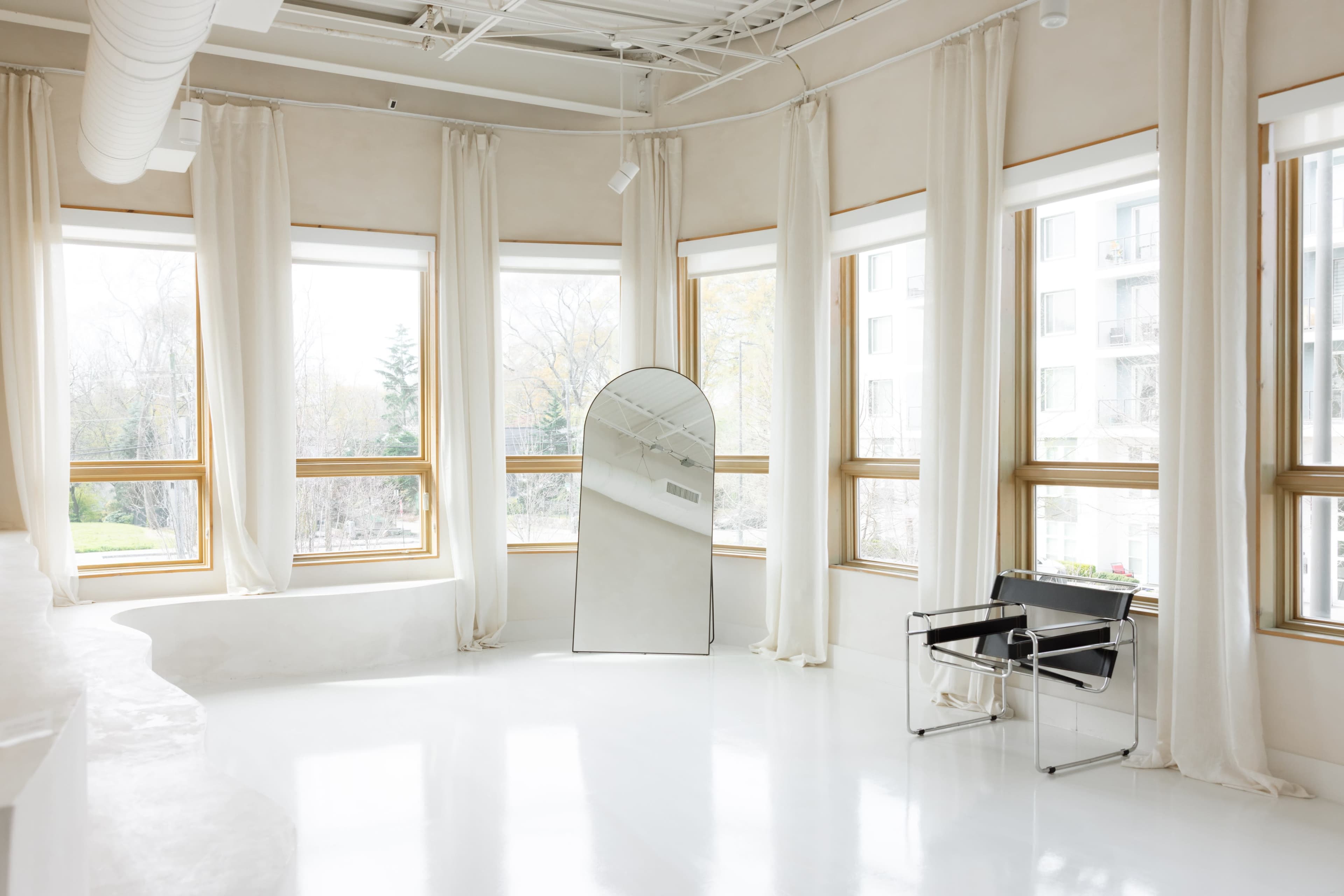 The image features a bright interior space with large windows, sheer curtains, a modern chair, and a curved mirror.