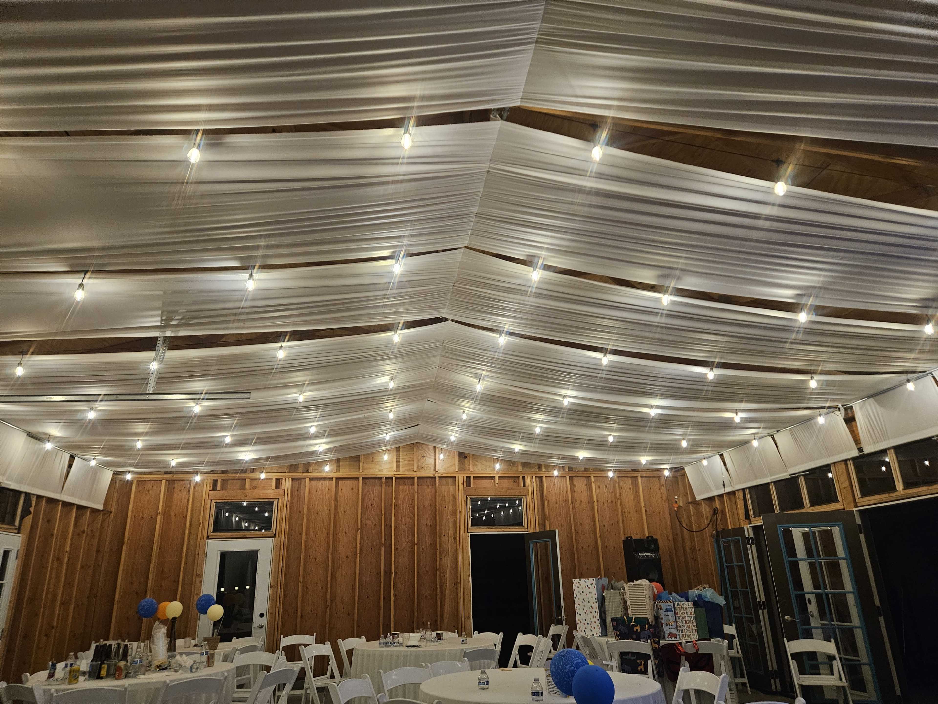 The image shows a banquet hall with draped white fabric ceiling and string lights, furnished with round tables and chairs.