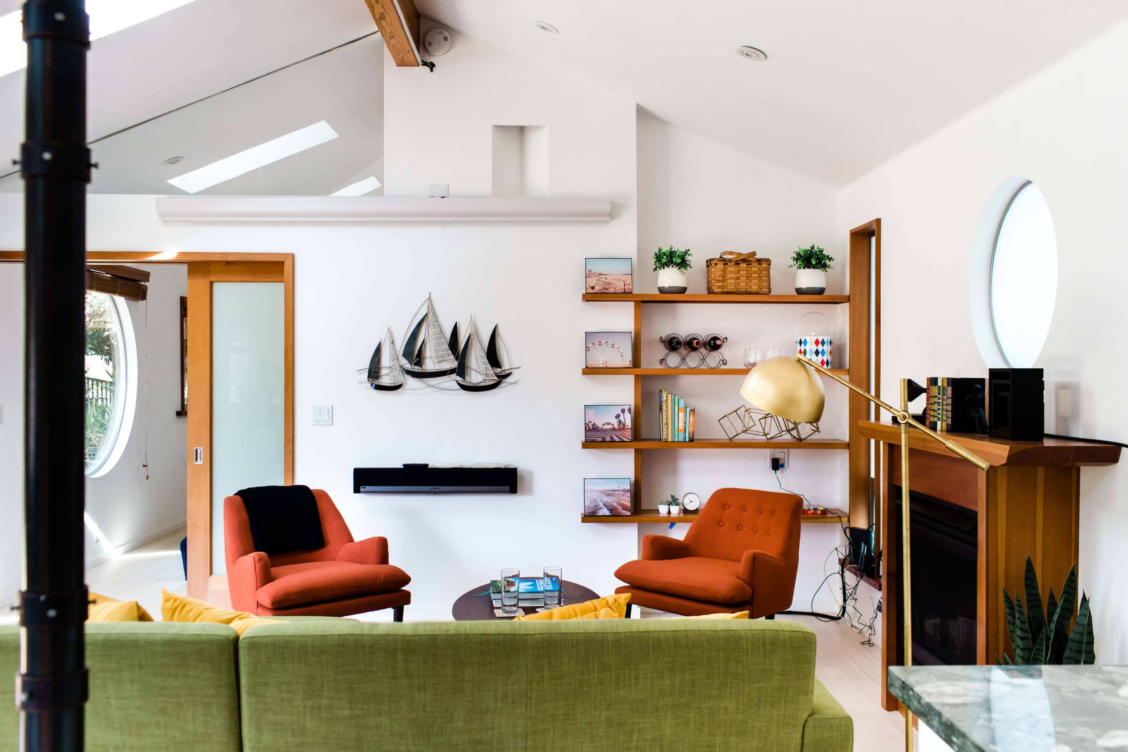 A modern living room features a green sofa, two orange chairs, a round coffee table, and shelves with decorative items and books.