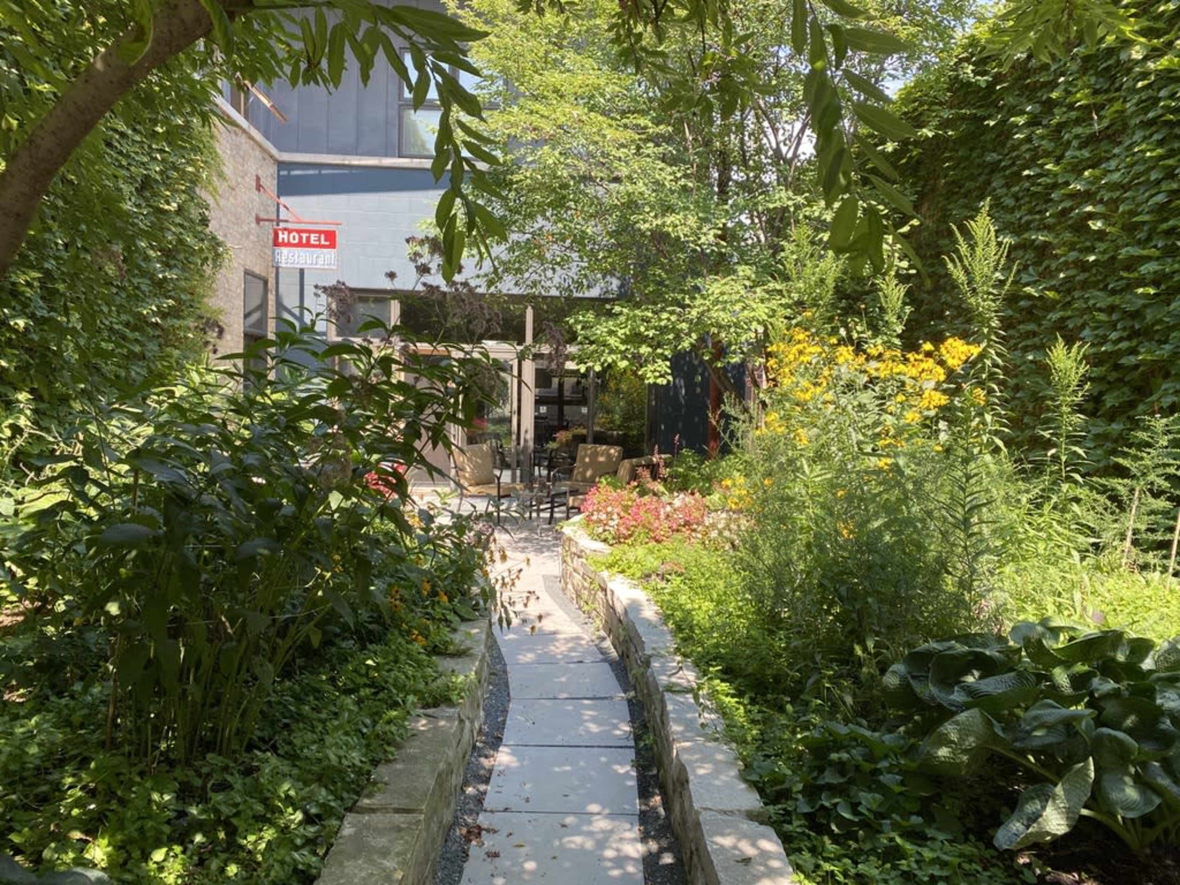 A narrow stone path leads through a lush garden filled with various plants and flowers, with a seating area visible in the background.