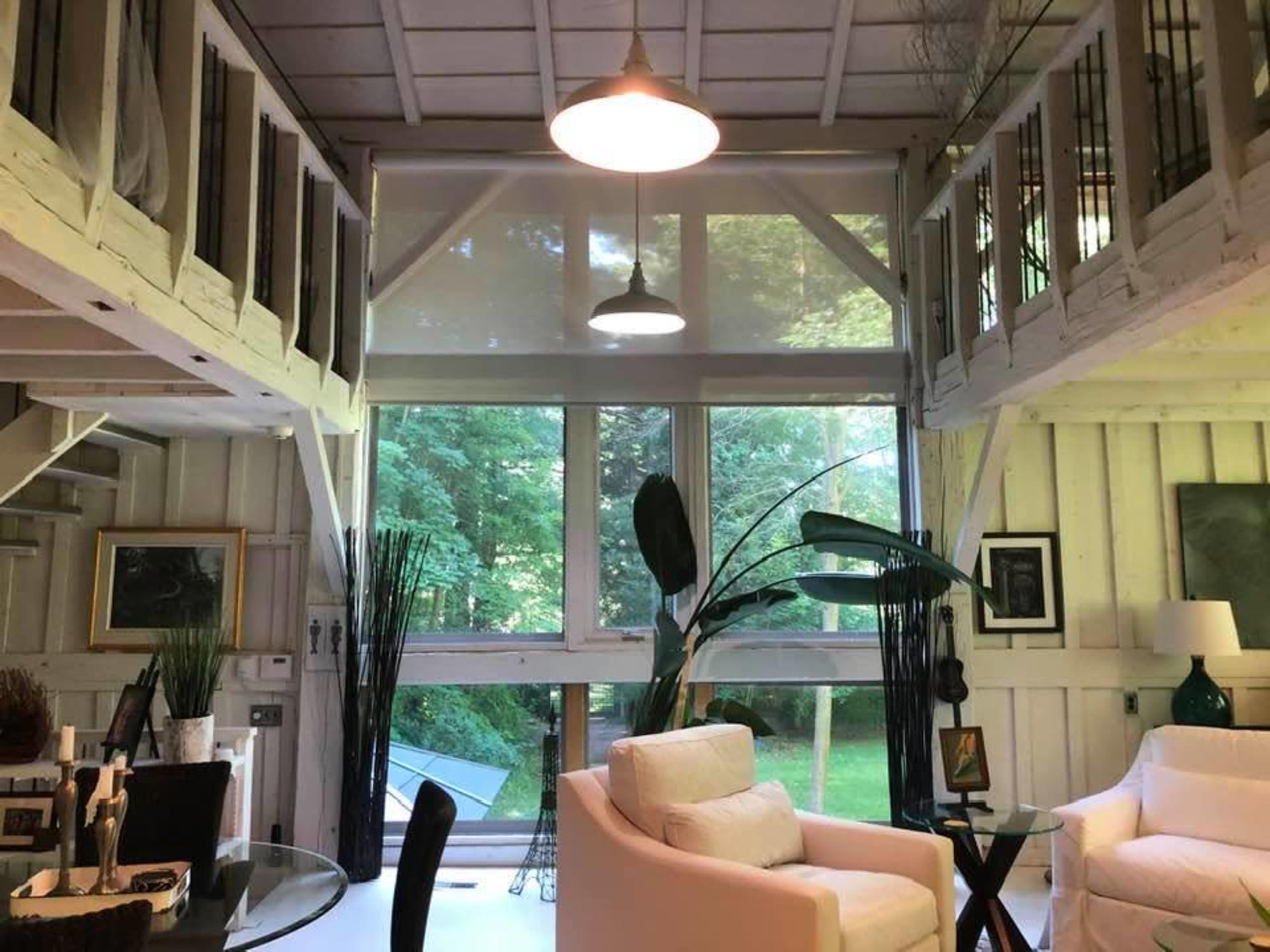 A bright interior of a modern loft-style living space with large windows, white furnishings, and a view of green foliage outside.